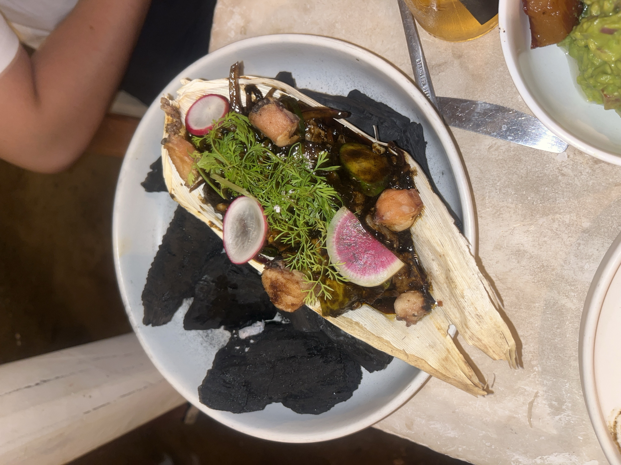 The image shows a partially eaten dish on a white plate. The dish consists of corn husks, dark sauce, small pieces of meat, radish slices, and green sprigs, arranged on top of charcoal fragments. The plate is resting on a beige surface next to a knife, a beer bottle, and another plate containing food (guacamole and meat). An arm is visible in the upper left corner. The setting appears to be a restaurant. Based on the information given, the location is Brisas de Zicatela, Mexico.