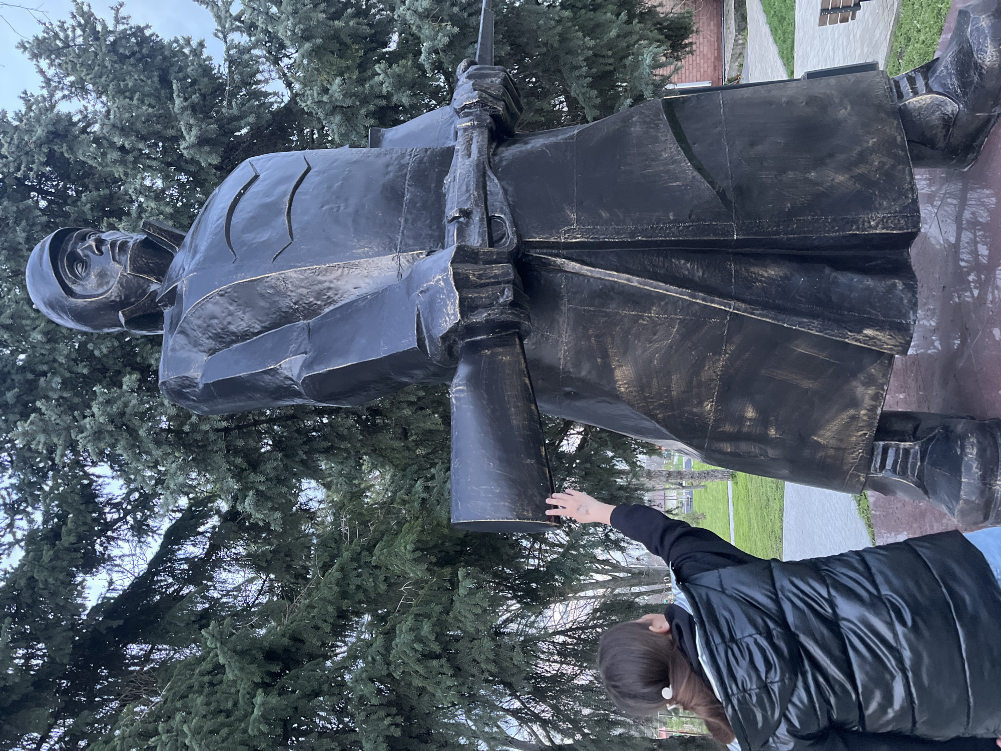 The image shows a large bronze statue of a soldier holding a rifle in a park-like setting. The statue is the main subject, dominating the frame with its dark, metallic finish. The soldier is wearing a helmet and a long coat. A woman stands near the statue, with her hand gently touching the rifle. The background features green trees, a walkway, and a glimpse of a bench. The location is likely the city of Novopokrovskaya, Russia.