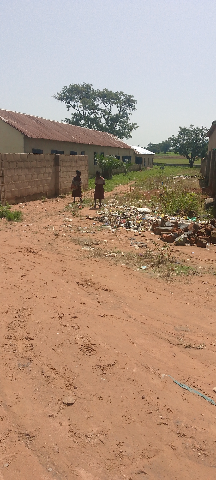 Two young females in traditional clothing stand in an outdoor, dusty environment near a brick wall and a building with a corrugated metal roof. The ground is covered in reddish-brown dirt, with scattered debris including plastic waste and bricks. There are tire tracks and footprints visible in the dirt. In the background, there are trees and another building. The sky is overcast and hazy.