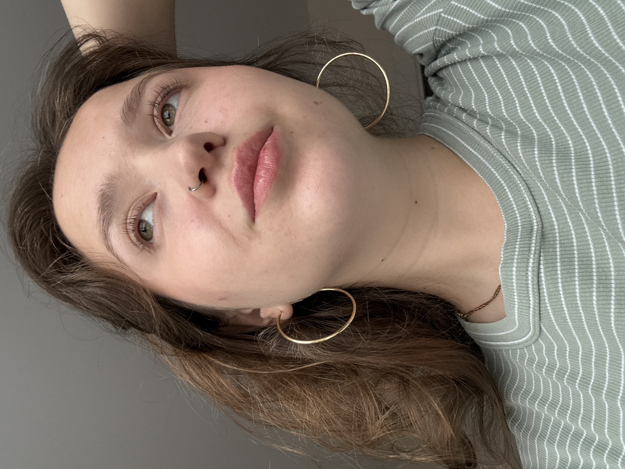 A young woman with fair skin, light brown hair, and green eyes is shown in a close-up, tilted portrait shot. She has a nose ring and is wearing large gold hoop earrings. Her lips are light pink. She is wearing a light green, striped shirt. A thin gold necklace is visible at her neck. The background is a solid gray color. The image does not contain any information about Izhevsk, Russia.