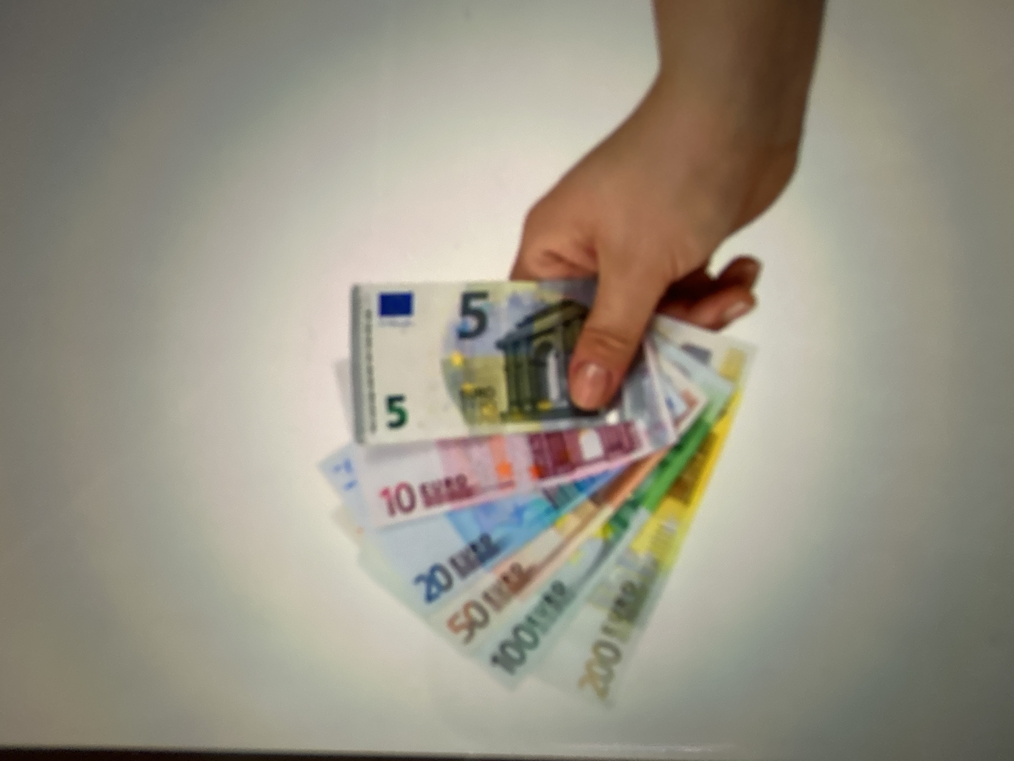The media file depicts a hand holding several euro banknotes fanned out. The subjects are a human hand and the banknotes, which are in denominations of 5, 10, 20, 50, 100, and 200 euros. The banknotes are colorful and appear to be real currency. The hand is holding them in a way that suggests they are being presented or displayed. The background is a plain, light-colored surface, possibly a table or wall, with a soft shadow cast by the hand and the money. The lighting is diffused, creating a gentle illumination. There are no discernible environmental cues that would indicate a specific time of day, weather, or location within Gelendzhik, Russia, other than the currency itself. The overall mood is neutral, with no strong emotions conveyed. The visible text includes the denomination numbers on each banknote, such as "5 EURO," "10 EURO," "20 EURO," "50 EURO," "100 EURO," and "200 EURO." The European Union flag symbol is also visible on the 5 euro note. The image is somewhat blurry, making precise details difficult to discern.