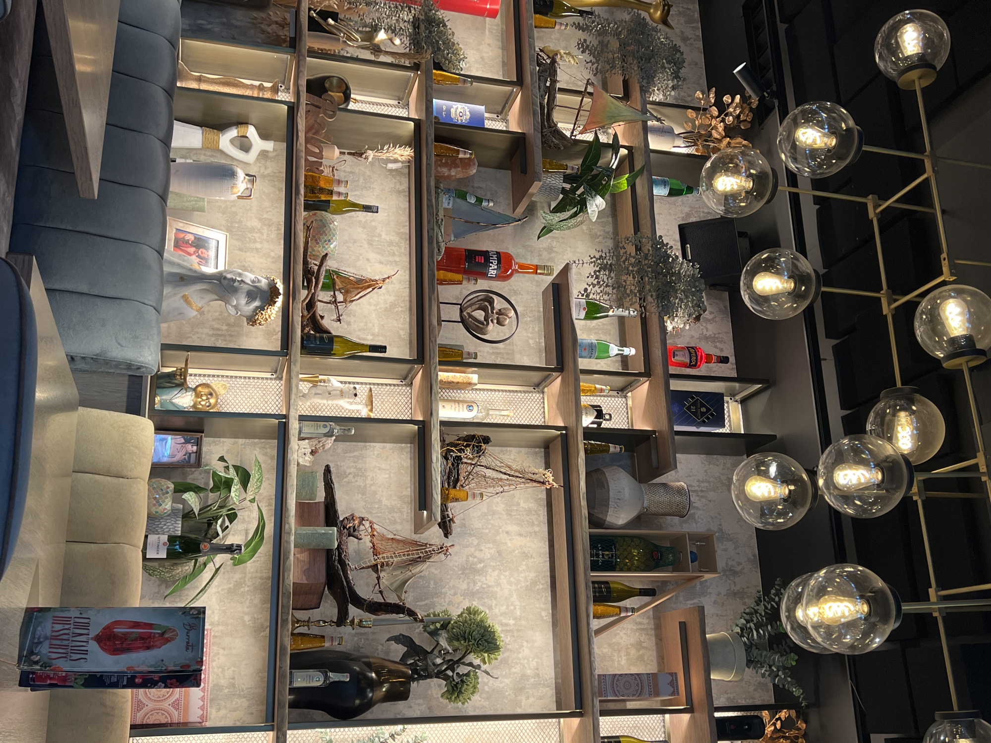 The image depicts the interior of a modern restaurant or bar in Sofia, Bulgaria. The scene is characterized by a stylish, contemporary design with exposed brick-like walls and industrial-style pendant lighting. Shelves are filled with various decorative items, including bottles of alcohol, small model sailboats, vases, plants, and decorative objects. The lighting is atmospheric, with several Edison-style bulbs creating a warm glow. In the foreground, part of a booth seating area is visible, with upholstered seating and wooden tables. The overall impression is one of a trendy establishment aiming for a sophisticated yet relaxed ambiance.