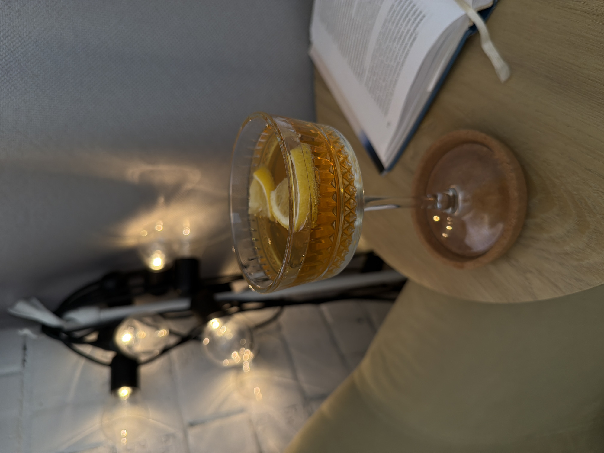 The image depicts a cocktail glass filled with a yellow liquid and two lemon slices, sitting on a wooden coaster. A partially open book with a blue cover and white bookmark is positioned next to the glass. In the background, a string of lights is draped over a possible ledge or surface. The scene appears to be indoors. The location and city are not discernible from the image.