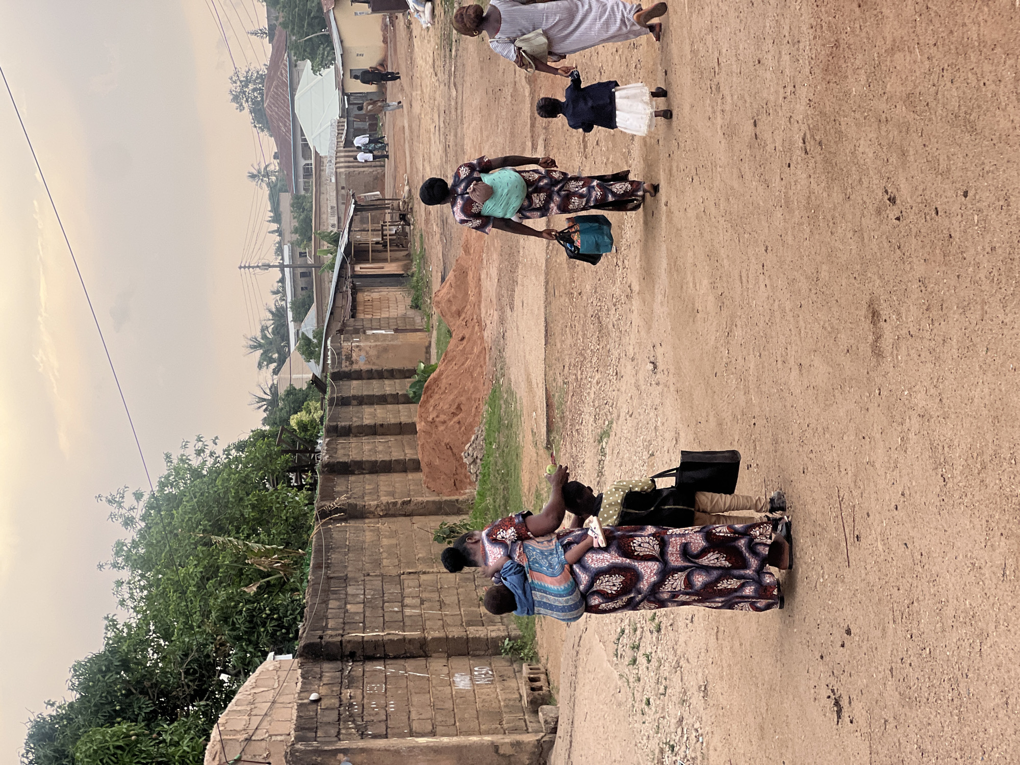 The image shows a street scene in Gura Top, Nigeria. The street is unpaved, with sandy soil. There are buildings on both sides of the street, constructed with brick walls and corrugated iron roofs. The buildings appear to be residential homes. Vegetation includes trees and grass growing near the buildings.
Several individuals are walking along the street. One woman is carrying a baby on her back and holding a bag. Another woman is holding the hand of a child who is walking next to her. In the distance, there are several more individuals walking along the street. The sky is overcast.