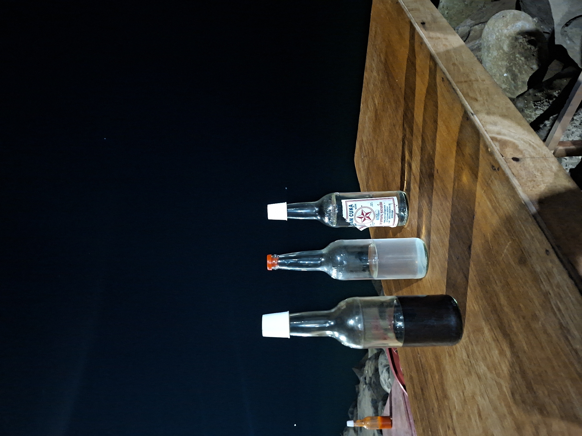 The scene depicts three glass bottles, each capped, lying horizontally on a wooden surface. The bottles vary in content: one appears to contain a dark liquid, another clear liquid, and the third has a label indicating it is “AM CUKA.” In the background is a dark sky or night scene, suggesting an outdoor setting. Based on the context provided, the location is likely in Lhokseumawe, Indonesia. The bottles are lined up, and the wooden surface is illuminated, possibly by artificial light. There are also stones in the background. There are no people visible.