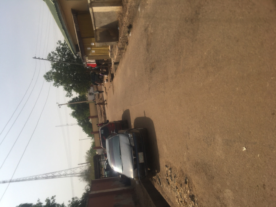 The image depicts a daytime street scene in Pindiga, Nigeria. The foreground shows a reddish-brown, unpaved or poorly paved road, with a shallow, concrete drainage ditch running along its left edge. Two passenger cars are parked or stationary on the left side of the road: a dark-colored sedan with visible front bumper damage is in the immediate foreground, followed by a reddish-brown sedan.

On the right side of the road, a single-story commercial building features a light green corrugated metal roof and light brown walls. A wooden storefront section displays the word "PHOTOCOPY" in dark lettering. A low concrete wall separates the building from the road. Several individuals are present: approximately six adults are visible in the midground, walking or standing on the road and near the building on the right, some appearing to interact. Another individual is partially obscured behind the dark sedan on the left.

The background on the left features a reddish-brown perimeter wall with foliage behind it. A very tall, slender metal lattice communications tower dominates the left background, with another smaller tower visible further back. Multiple utility poles with overhead power lines span across the scene. The sky is clear and bright, indicating daylight conditions.
