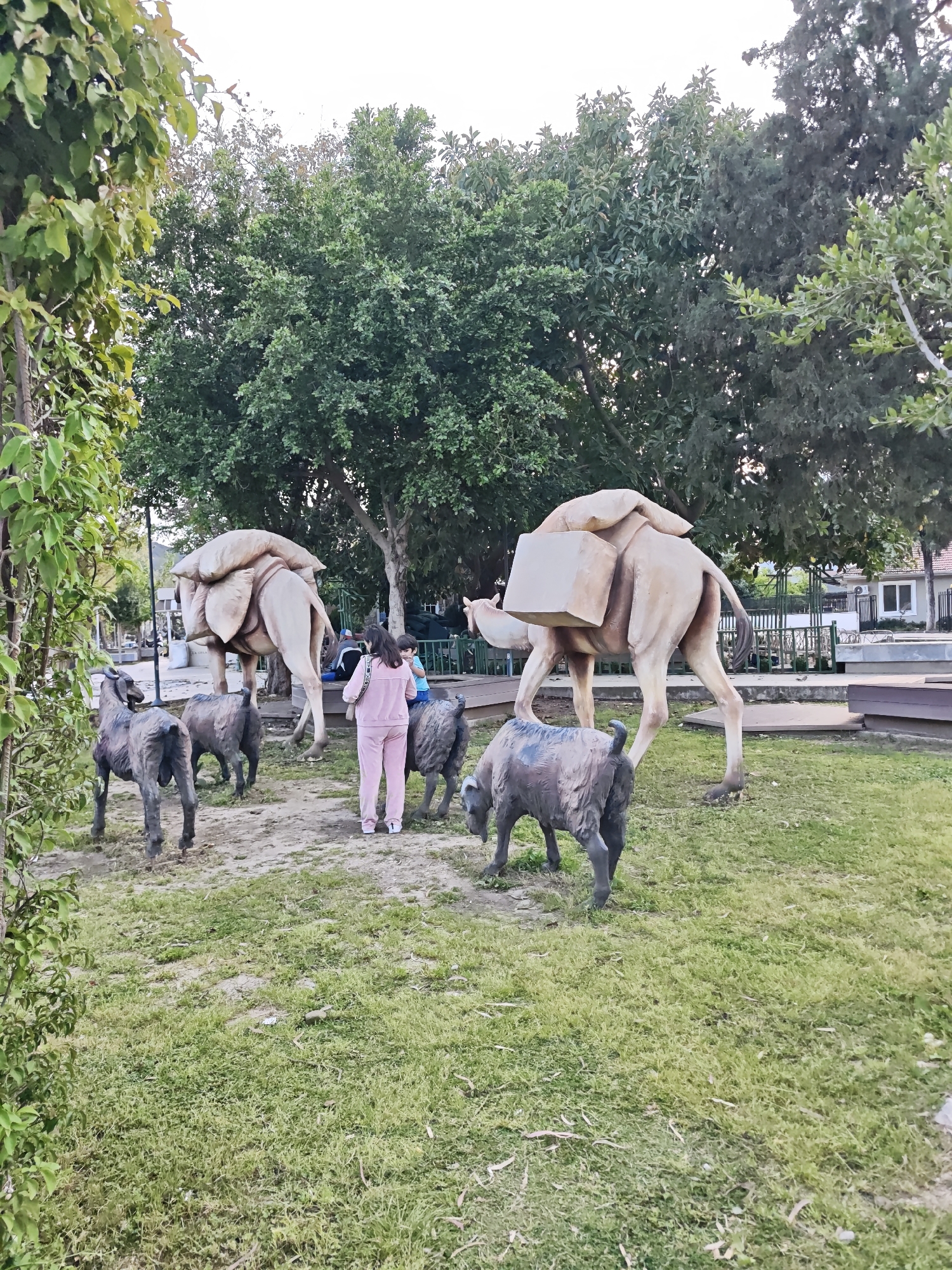 The image shows a public park in the city of Kemer, Turkey. In the park are several statues depicting camels and smaller ruminant animals. Two larger statues of camels, styled as pack animals with burdens on their backs, stand on a grassy area. Several smaller statues, resembling goats or small buffalo, are also present, scattered around the camels. A woman wearing a pink tracksuit is standing among the statues, partially turned away from the camera, with two children nearby. The park is landscaped with trees and grass. In the background, a structure with a metal railing is visible, along with some residential buildings. The sky is overcast.