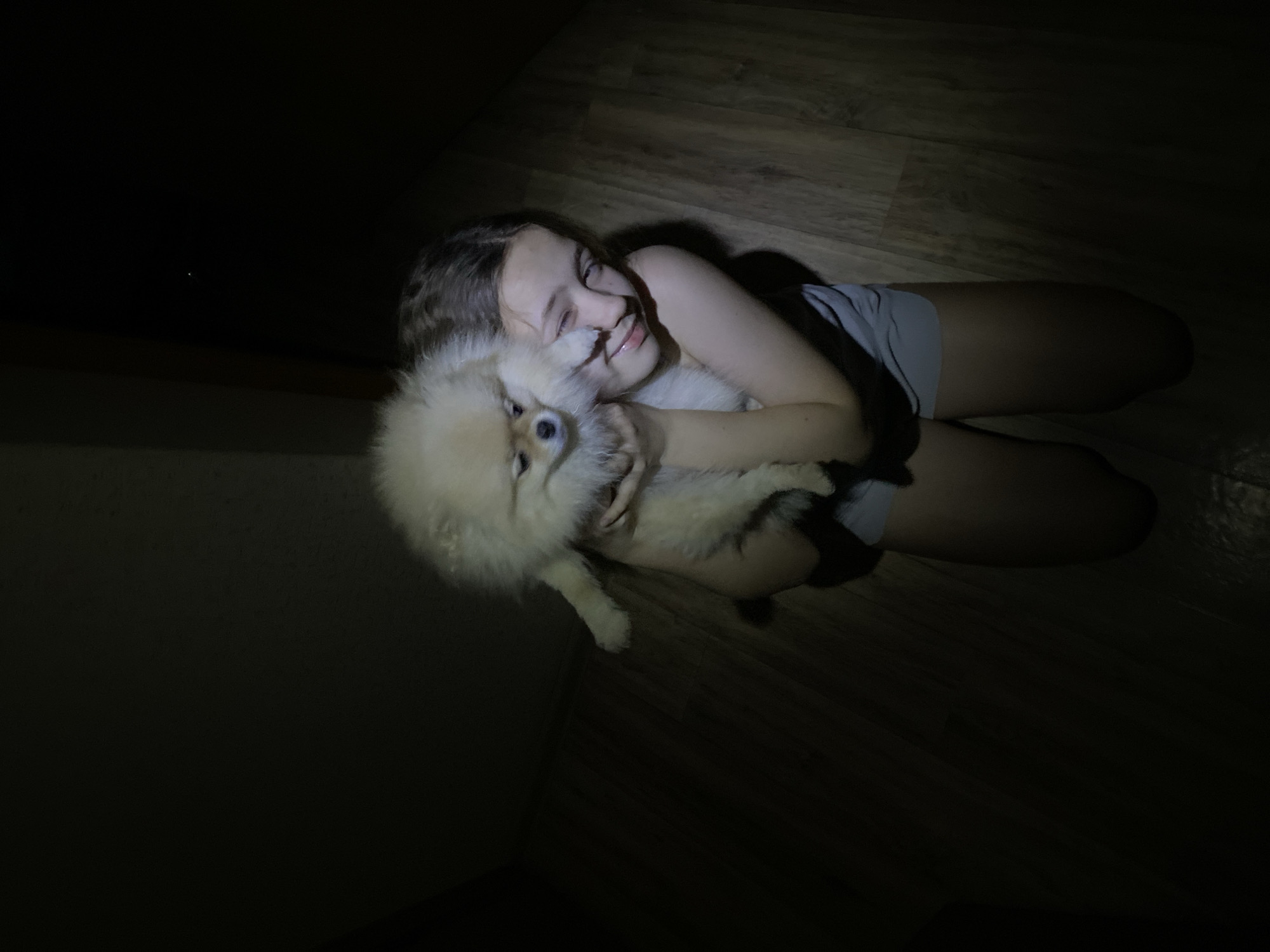 A girl is lying on a wood-patterned floor, hugging a small, fluffy dog. The room is dimly lit, with the primary light source illuminating the girl and the dog. The girl is wearing a grey shirt and appears to be smiling. The dog is white and fluffy and looks to be a Pomeranian. The scene is situated indoors.