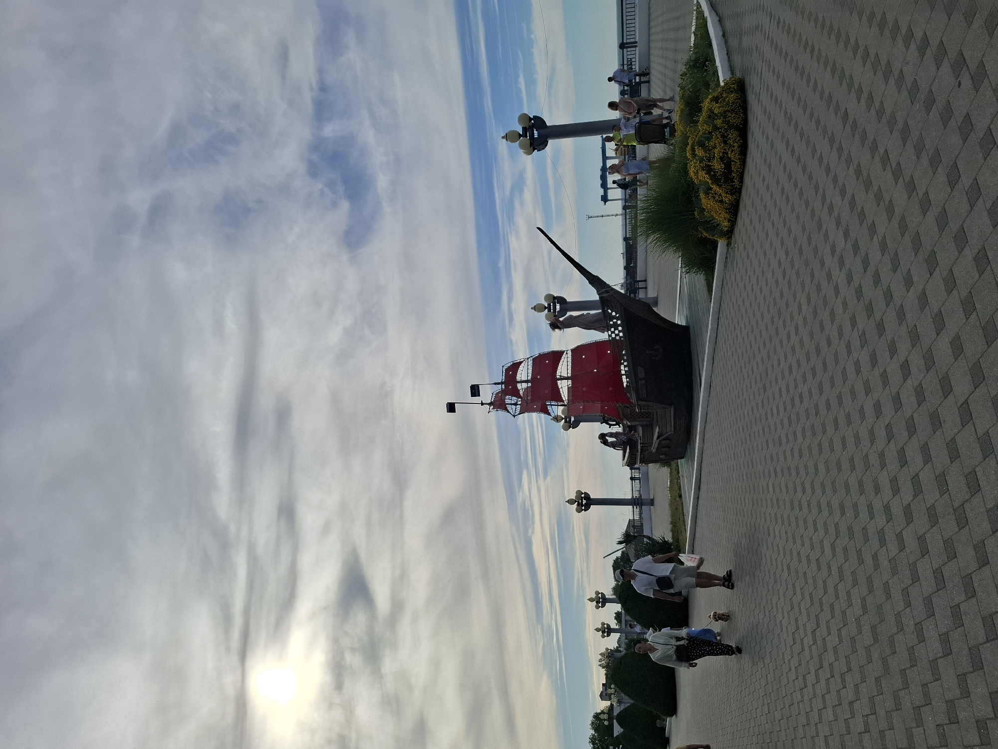 An outdoor, eye-level, medium-wide shot captures a public promenade with a decorative pirate ship replica in the background under a partly cloudy sky. The sun is visible in the upper left corner, casting a bright glare. In the foreground, a textured grey paved walkway is prominent. Two people are walking on the walkway; a woman in a light-colored shirt and dark polka-dot pants, accompanied by a small, light-brown dog on a leash, and a man in a white shirt, beige shorts, and a white cap, carrying a white bag. Behind them, several large, dark green, cone-shaped bushes line the left side of the promenade.
The pirate ship replica, the central focus of the middle ground, is a dark-colored vessel with bright red sails and black flags. It is positioned on a raised area with some people visible near it. Several tall lampposts with multiple lights are spaced along the promenade. In the background, beyond the ship, a railing runs along the edge of the promenade, with a view of what appears to be the sea or a large body of water. More people are scattered on the promenade in the distance, some sitting on benches. The overall scene suggests a public recreational area, possibly a waterfront, on a bright, partly cloudy day.