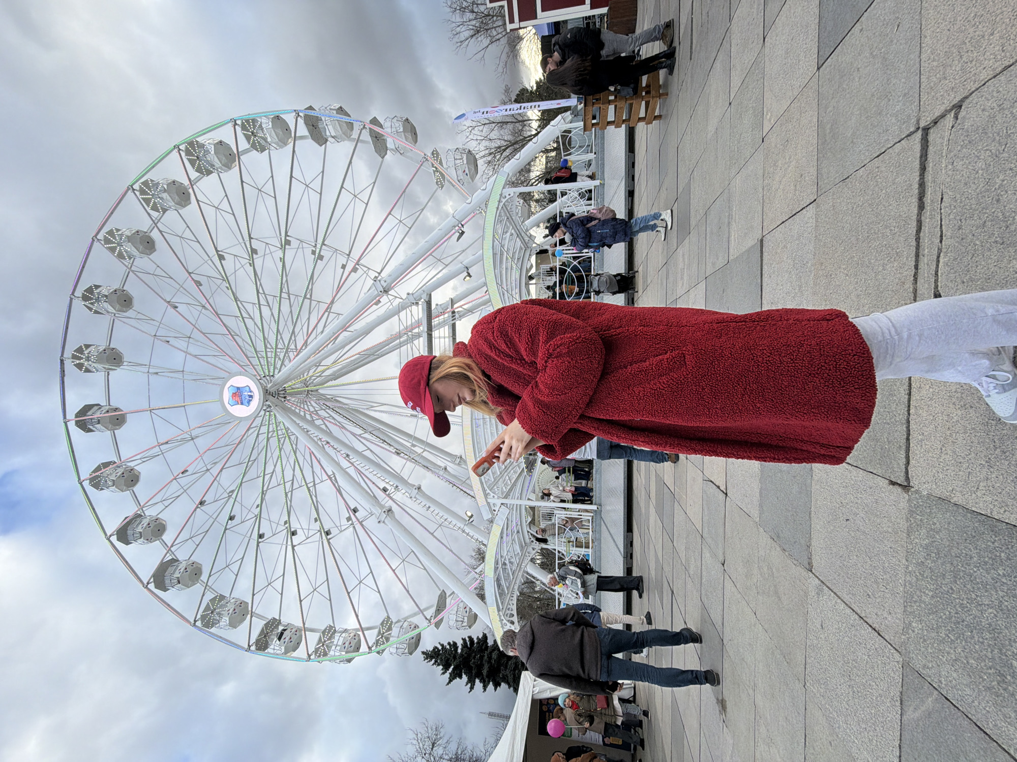 A medium, full shot captures a young woman, presumed to be in her early twenties, engrossed in her phone. She's wearing a red cap and a plush red coat, complemented by light grey sweatpants and white sneakers. She's positioned to the right of a large Ferris wheel. The wheel has grey passenger cars and is adorned with colored lights. The sky behind the Ferris wheel is overcast and grey. To the right of the woman, there are several people standing and sitting along a white railing. In the foreground, directly in front of the wheel, there's a man in a blue sweater and jeans. In the lower left corner, there's a Christmas tree. The ground is paved with grey stone tiles. The overall setting suggests an amusement park or festival located in a city. Based on the distinctive Ferris wheel and the surrounding architecture, this is most likely the Christmas market located in Sofia, Bulgaria.