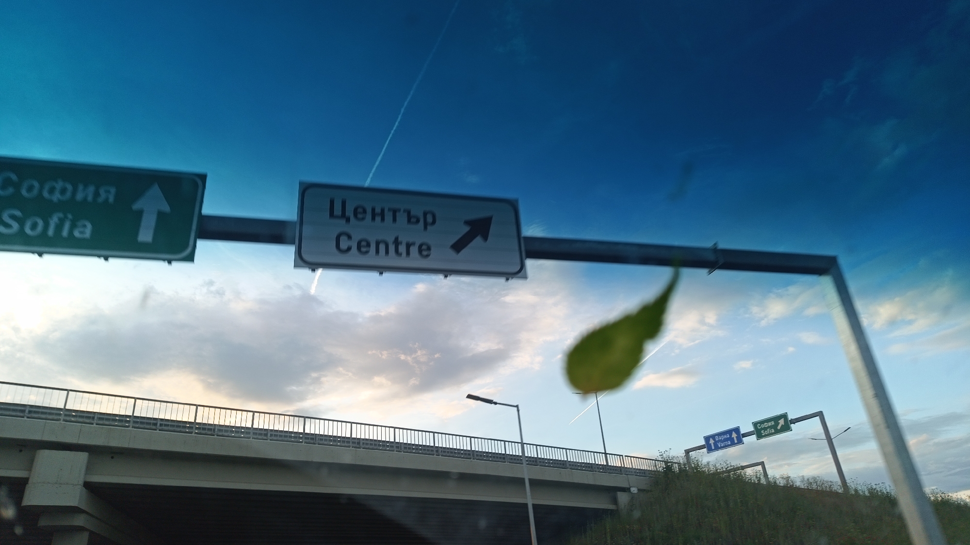The image depicts road signs above an overpass. One green sign points upward to Sofia. A white sign to the right points toward the "Centre" of the city. In the lower right of the image is a blue sign for Varna and another green sign pointing to Sofia. The overpass has a metal railing. A leaf is partially obstructing the center of the frame. The sky is blue with light clouds and contrails. Based on the road signs present, the location is likely in or around Burgas, Bulgaria.