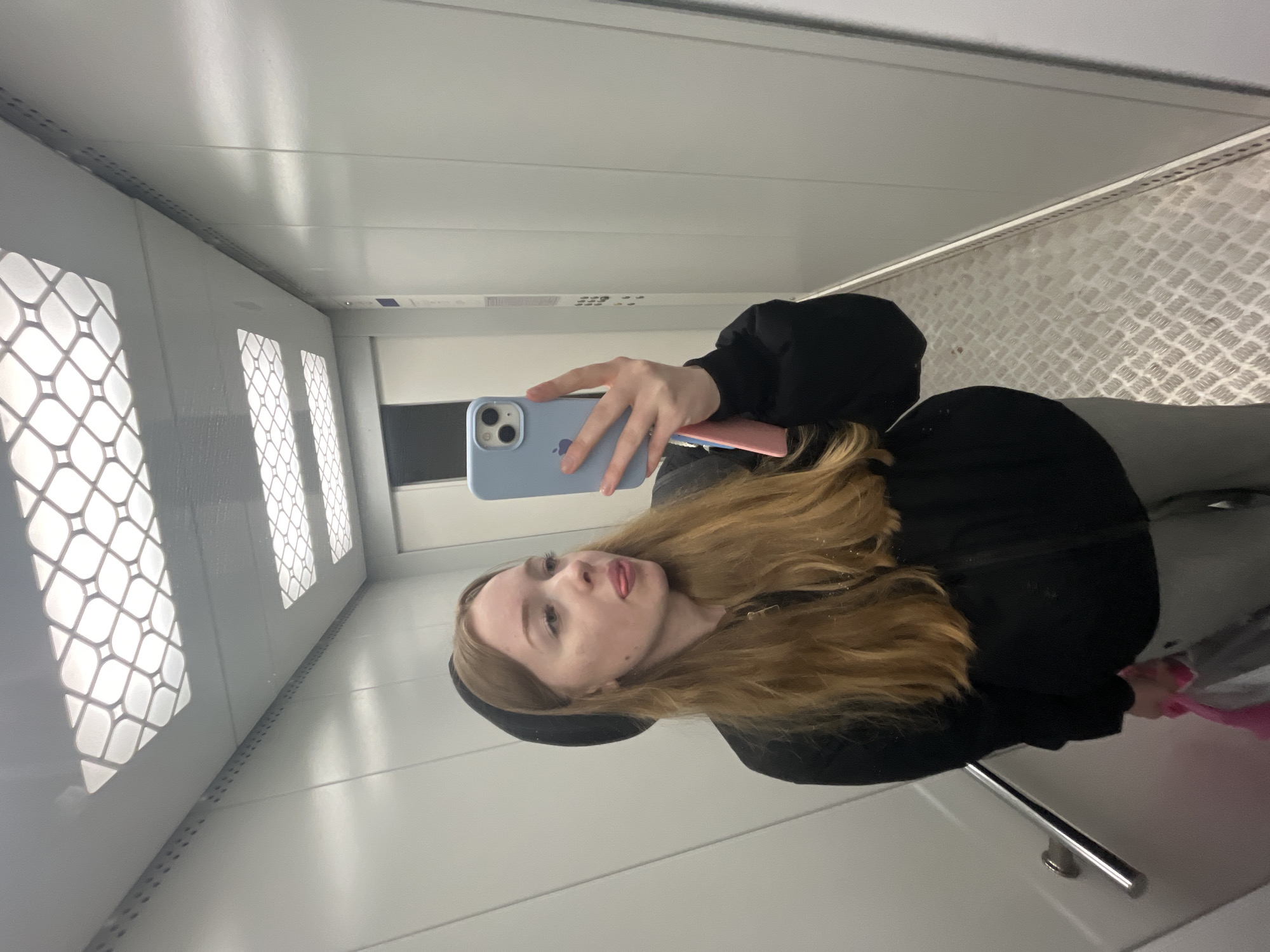 A young woman captures a mirror selfie inside what appears to be an elevator or a small, enclosed room. The image is rotated, showing her lying horizontally, but she is posing upright. She is seen from the chest up, with long, light brown hair flowing over her shoulders, wearing a black long-sleeved top or hoodie and a black beanie. Her expression is playful, with her tongue slightly out as she looks directly into the camera.
She holds a light blue iPhone, featuring a visible Apple logo, in her right hand. A small pink object, possibly another phone or case, is partially visible behind it. The setting's interior consists of light-colored, possibly white or metallic, paneled walls and ceiling. The wall to her left displays several rectangular sections with a decorative, diamond-grid pattern, which might be grates for ventilation or diffused lighting. The wall to her right features a textured, raised diamond-plate pattern, commonly found in elevators or industrial settings. A silver handrail is visible in the lower right of the frame. The space is brightly illuminated by artificial lighting, reflecting off the smooth and textured surfaces. The photo was taken in Chaikovsky, Russia.