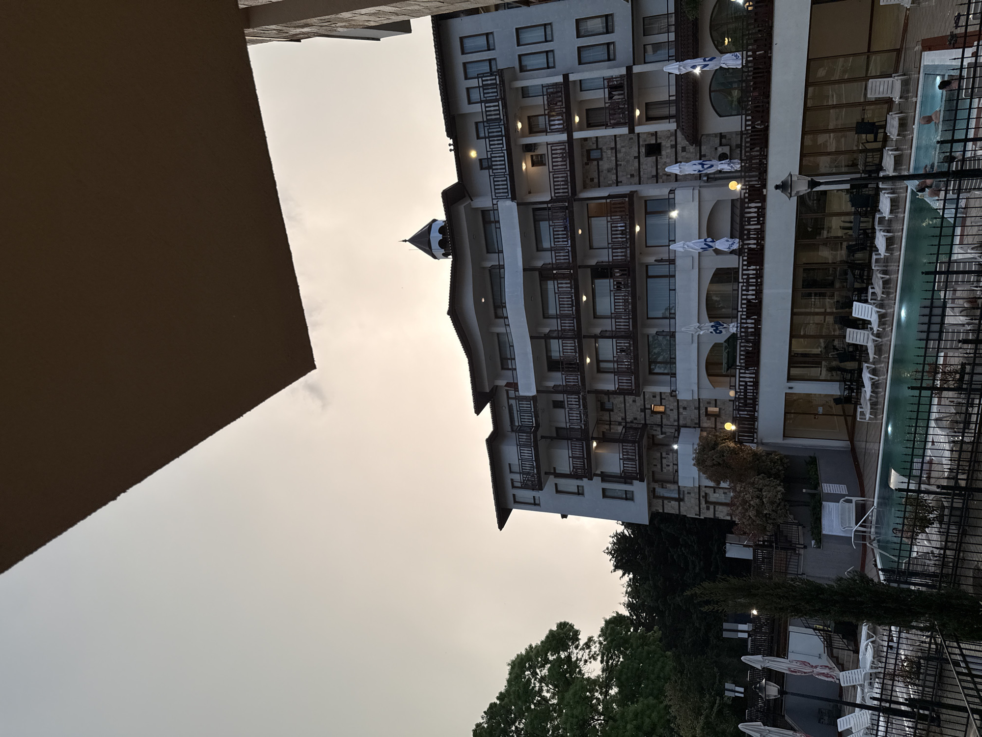 The image depicts a multi-story hotel in Varna, Bulgaria, observed from a lower vantage point. The building features a white facade with dark-framed windows and balconies with wooden railings. Several parasols are seen on the side of the hotel facing a swimming pool containing several persons. The background includes trees and a light overcast sky. A portion of a building is visible on the left side of the frame.