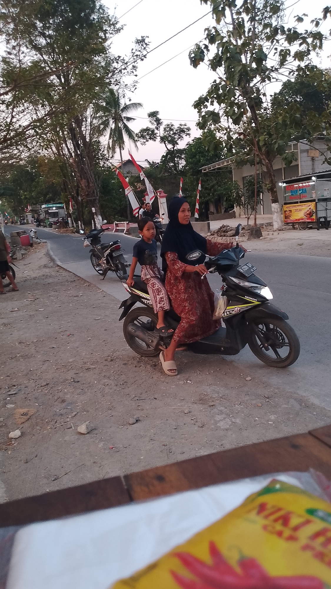 In Karanglangu, Indonesia, a woman wearing a black hijab and a patterned red dress rides a black motorcycle. A young boy in a dark blue shirt and patterned pants sits behind her, facing forward. They are on a paved road with a dirt shoulder. In the background, there are trees, buildings, and several Indonesian flags. The sky is overcast. A portion of a yellow package with red chili peppers is visible in the foreground.
