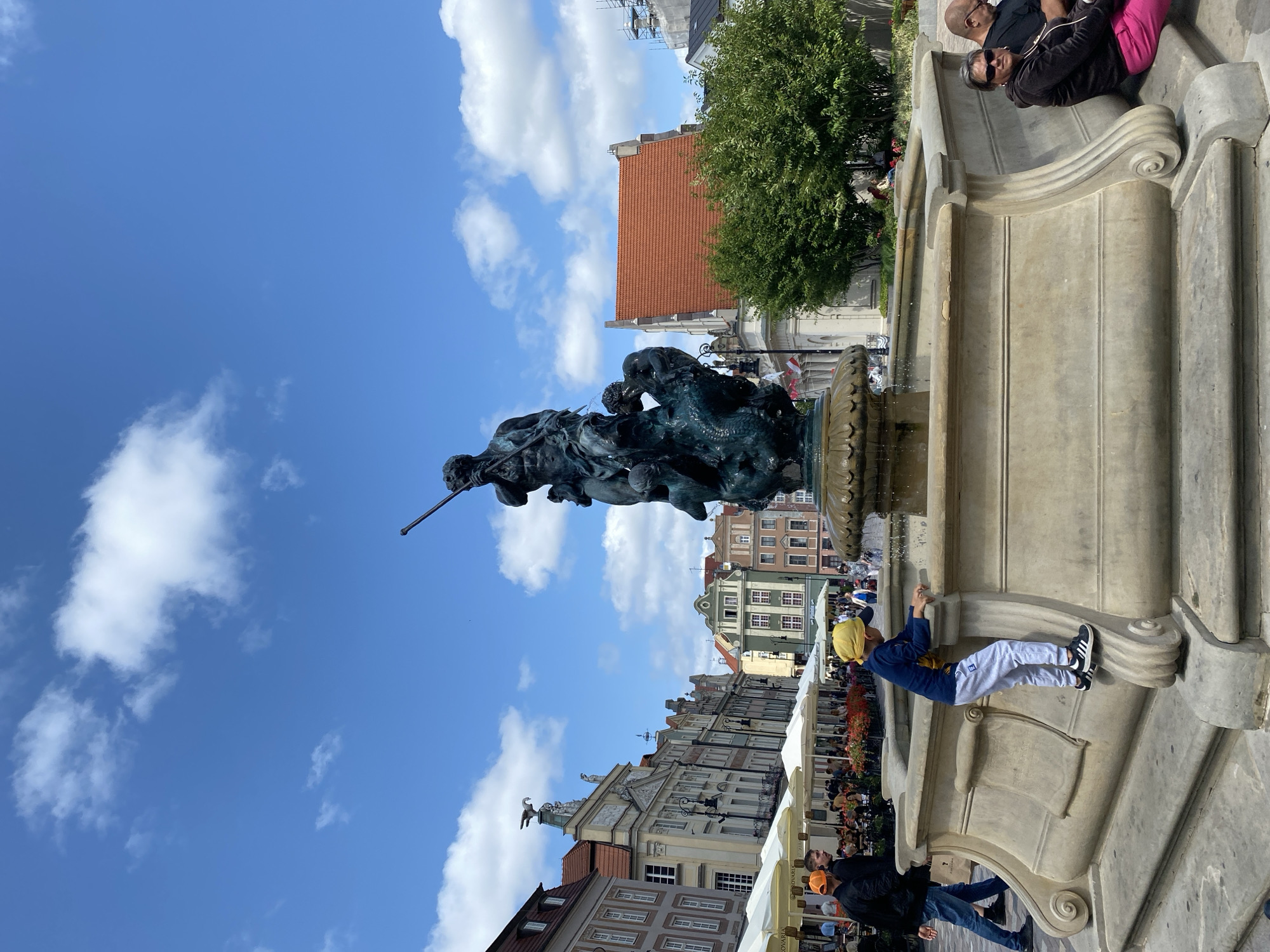 In Poznań, Poland, a young boy wearing a yellow hat, blue long-sleeved shirt, and grey sweatpants is climbing on the edge of a stone fountain. The fountain features a dark bronze sculpture of Neptune riding a sea creature, holding a trident. The fountain is situated in a public square with historic buildings lining one side. The buildings have colorful facades and traditional architecture. Umbrellas from outdoor cafes are visible, with people seated at tables. Other people are walking in the background. The sky is blue with scattered white clouds.
