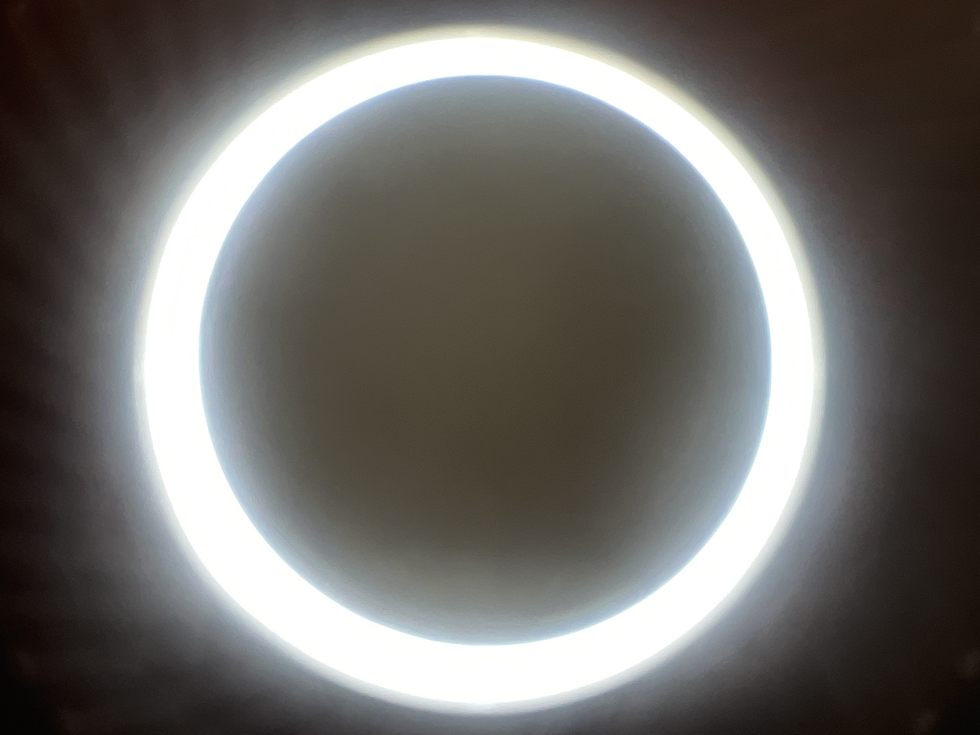 An abstract image features a bright, circular white light against a dark, muted background. The light forms a distinct ring, with the center of the circle appearing as a soft, diffused grey. The edges of the ring are sharp, indicating a direct light source, while the surrounding darkness suggests the light is contained within a space or viewed from a distance. The overall impression is one of glowing intensity, with a subtle gradient from the bright core to the darker periphery. The image's focus is entirely on the luminous ring, with no discernible details of people, objects, or a specific environment.