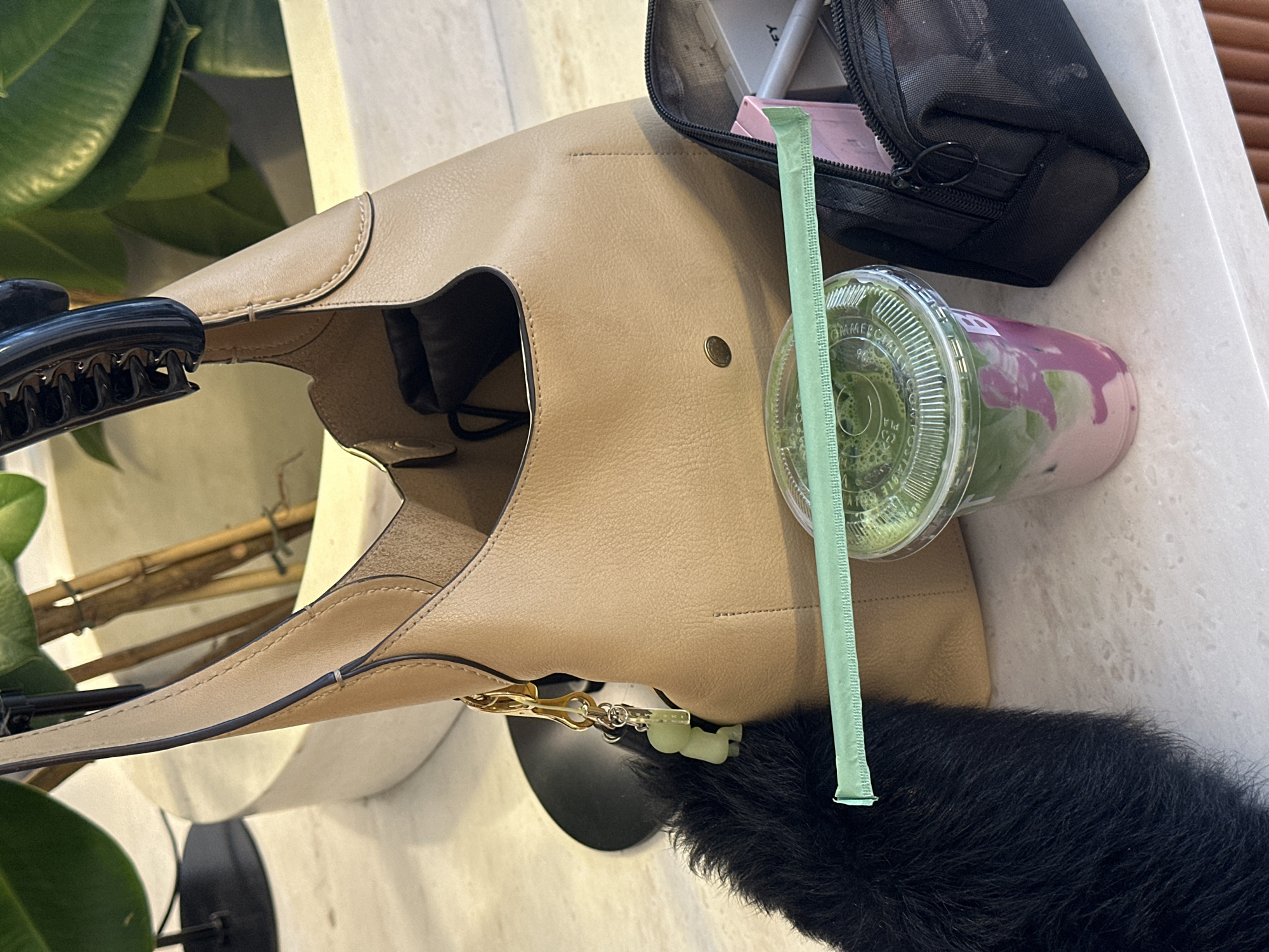 A close-up, eye-level shot depicts a casual arrangement on a surface. A tan leather hobo bag dominates the composition, positioned centrally. Next to the bag, a clear plastic cup containing a green beverage with a purple swirl rests. A green paper straw extends upwards from the drink. To the right of the bag, a black zippered pouch is visible. In the lower right corner, a portion of a dark, furry object is present. The background reveals a plant with large green leaves and thin brown branches. The scene is likely indoors, given the items present. The location cannot be determined from the image.