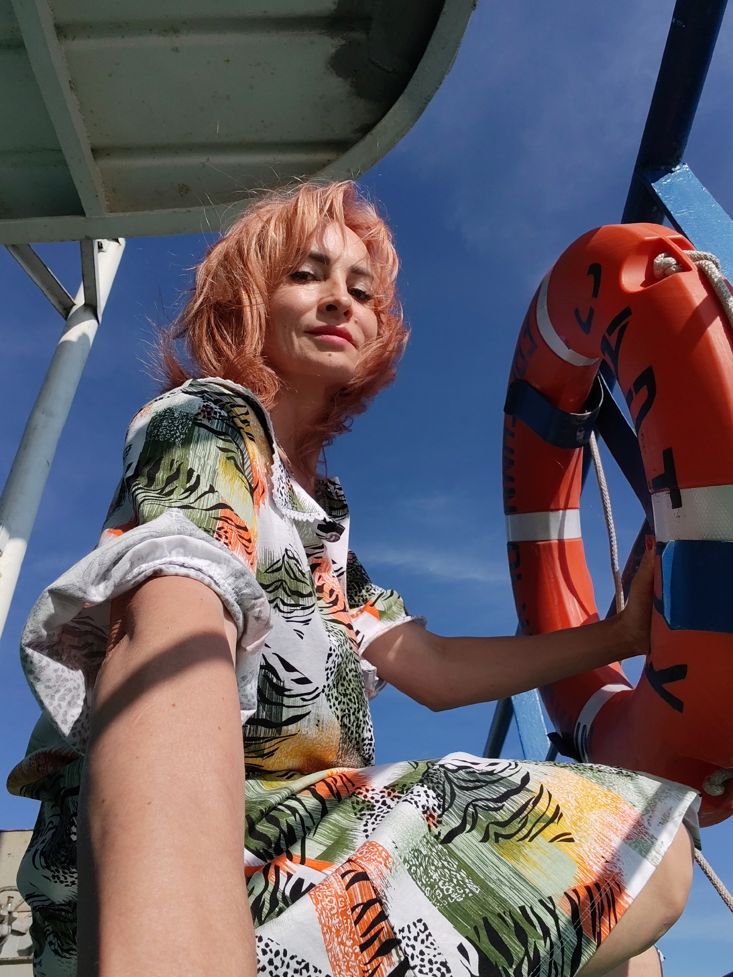 A woman with short, reddish-pink hair is sitting outdoors, possibly on a boat, as indicated by the life preserver and the structure above her. She is wearing a colorful, patterned dress with green, orange, and black abstract designs. Her left arm is extended towards the camera, and her right hand is resting on the orange life preserver. The background is a clear blue sky. The camera angle is from below, looking up at the woman. The location is not specified within Nova, Russia.