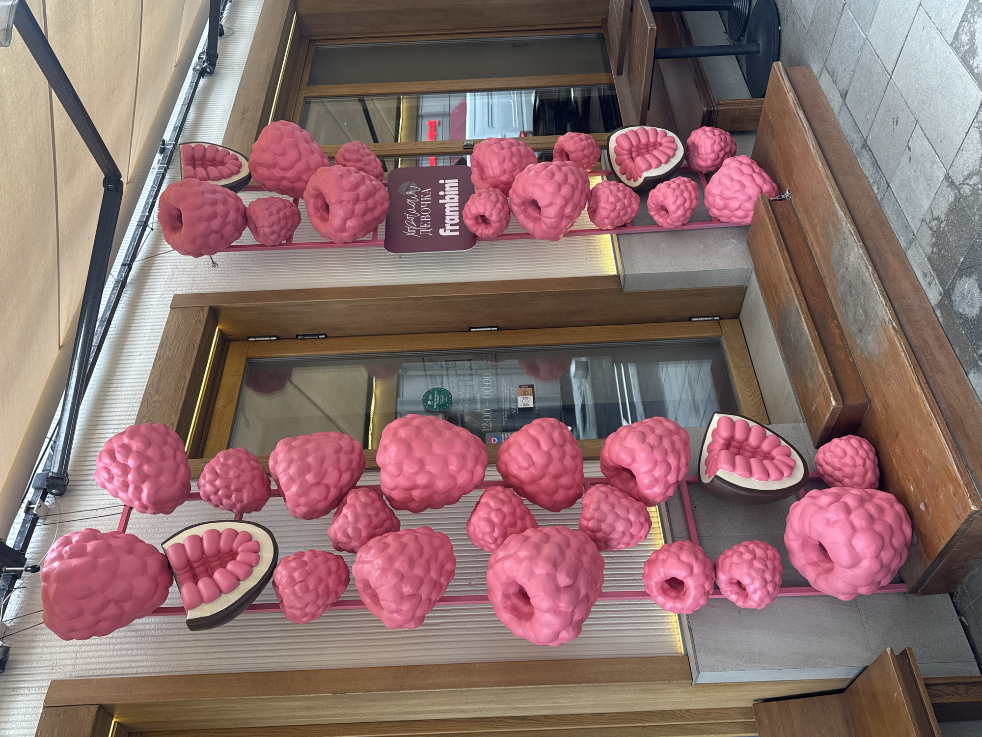 The image depicts the exterior of a building in Moscow, Russia. The building is adorned with a decorative display of oversized pink raspberry sculptures. The sculptures are of varying sizes and some are bisected to show the fruit's interior. They are mounted on a pink, rectangular frame attached to the facade between the windows. A sign with Cyrillic script and the name "frambini" is visible. The building has a wooden exterior around the windows. To the right of the display are wooden planks and a small portion of sidewalk is visible.