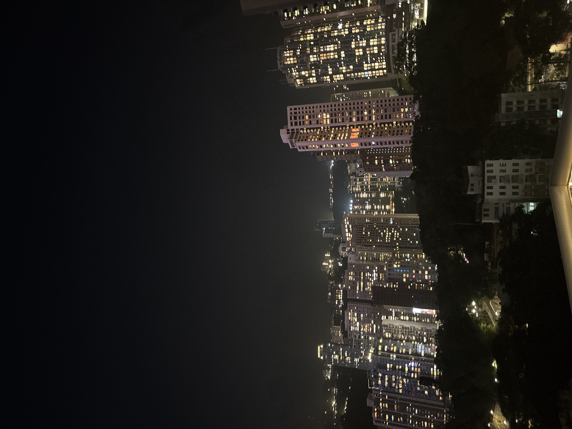The image captures a nighttime view of Hong Kong, China. On the right side of the frame are densely packed high-rise buildings with numerous illuminated windows, indicating residential or commercial activity. Patches of greenery interspersed among the buildings suggest urban parks or tree-lined streets. To the left, a large expanse of darkness dominates the scene, likely representing the night sky or a body of water adjacent to the city. A portion of a railing or structure is visible on the far right.