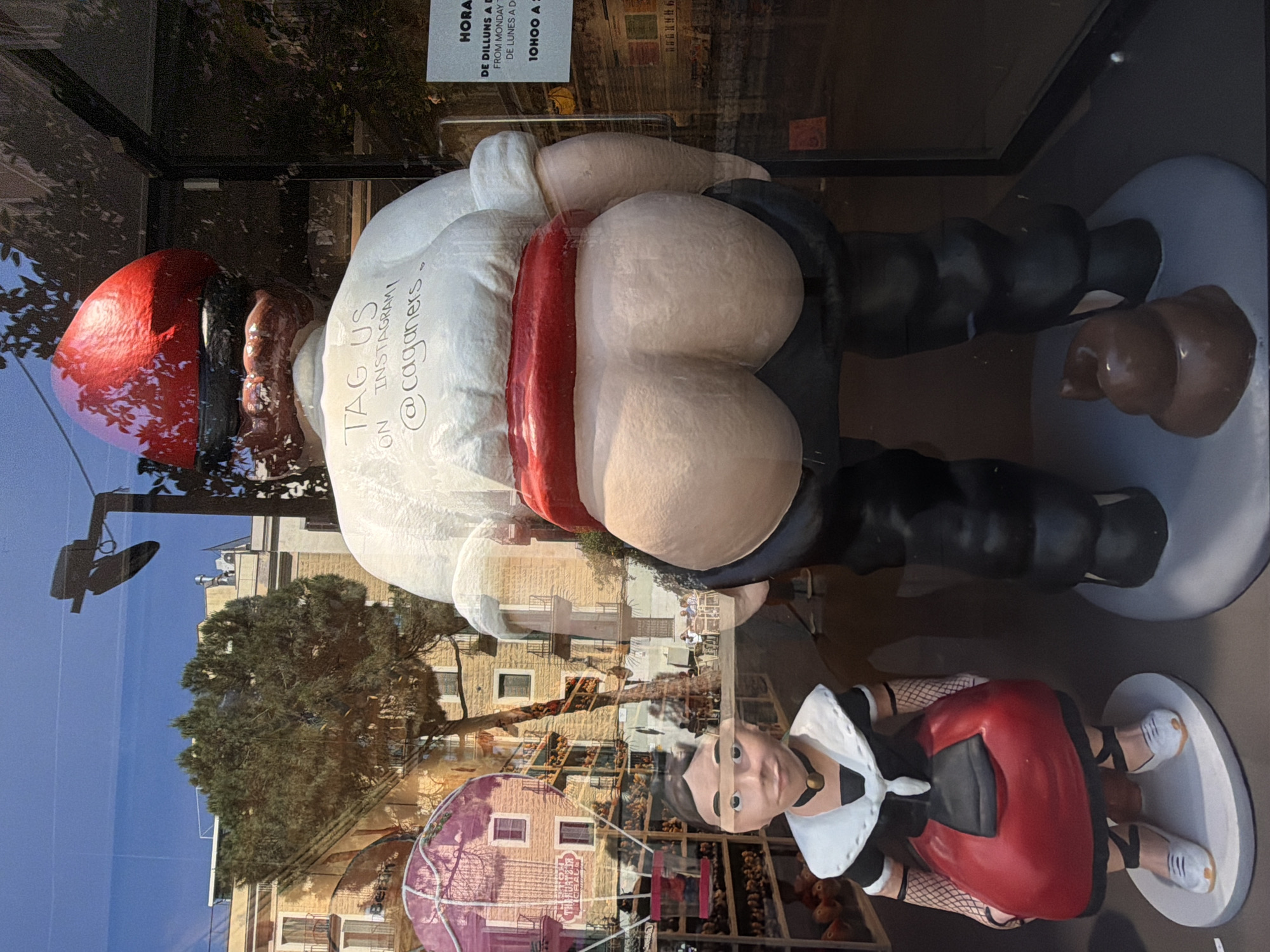 The image shows a display of caganers, traditional Catalan figurines that depict individuals defecating, in a store window. The figurines are on display in Barcelona, Spain, judging by the buildings reflected in the window. The primary figurine is Santa Claus, dressed in his typical red hat and suit, with his buttocks exposed and a pile of feces nearby. Another figurine appears to be a woman in a red and black dress. The window reflection shows buildings, trees, and a lamppost outside.