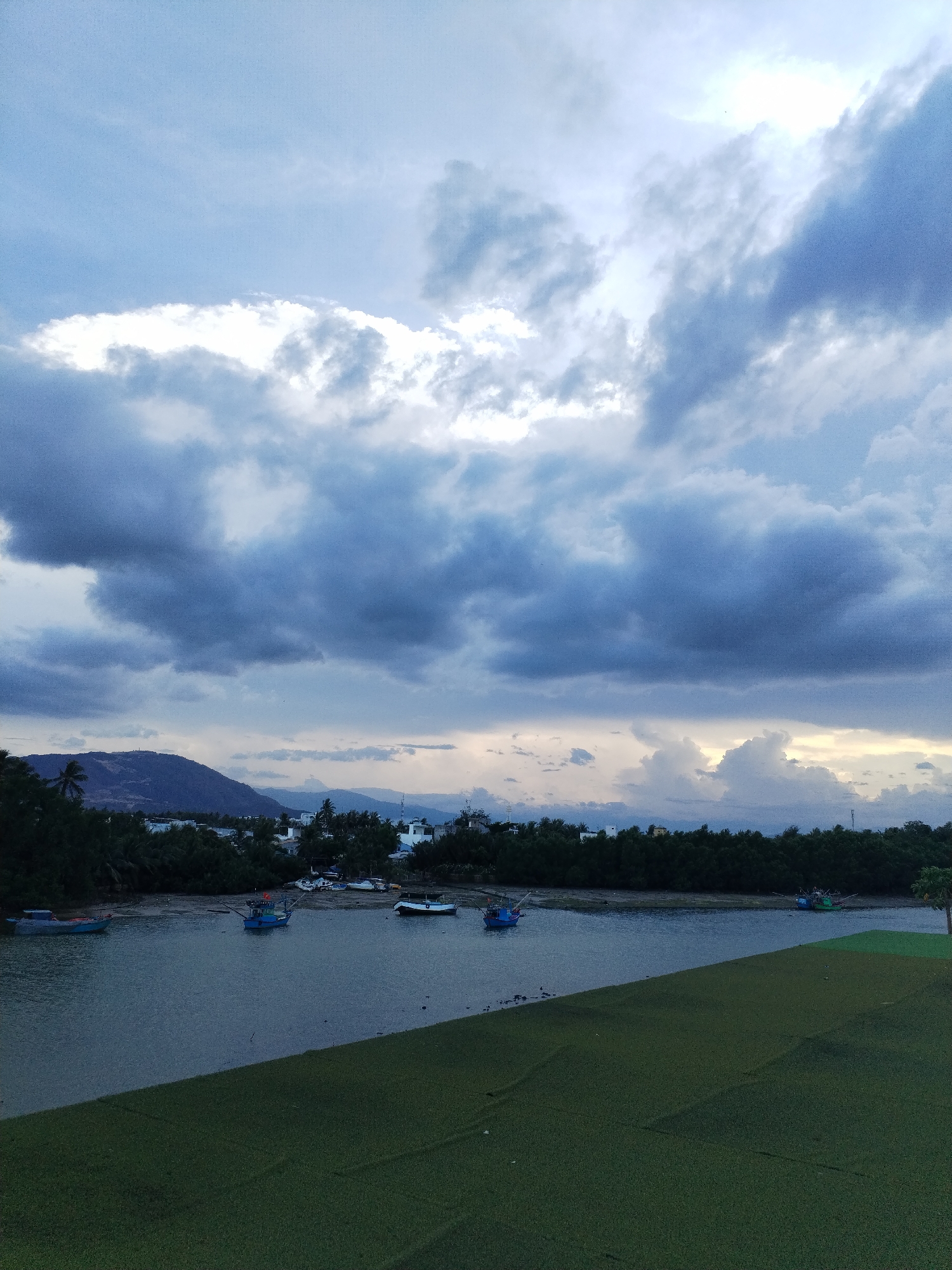 The image depicts a scene near Bac Nha Trang Ward, Vietnam, featuring a body of water with several small fishing boats anchored along the shore. The water is calm and reflects the overcast sky. A strip of green, possibly vegetation or grass, is visible in the foreground. The background reveals a shoreline lined with trees and buildings, suggesting a developed area. Distant mountains can be seen on the horizon, under a sky filled with dark, ominous clouds. The overall atmosphere is overcast and somewhat gloomy. No people are visible in the frame.
