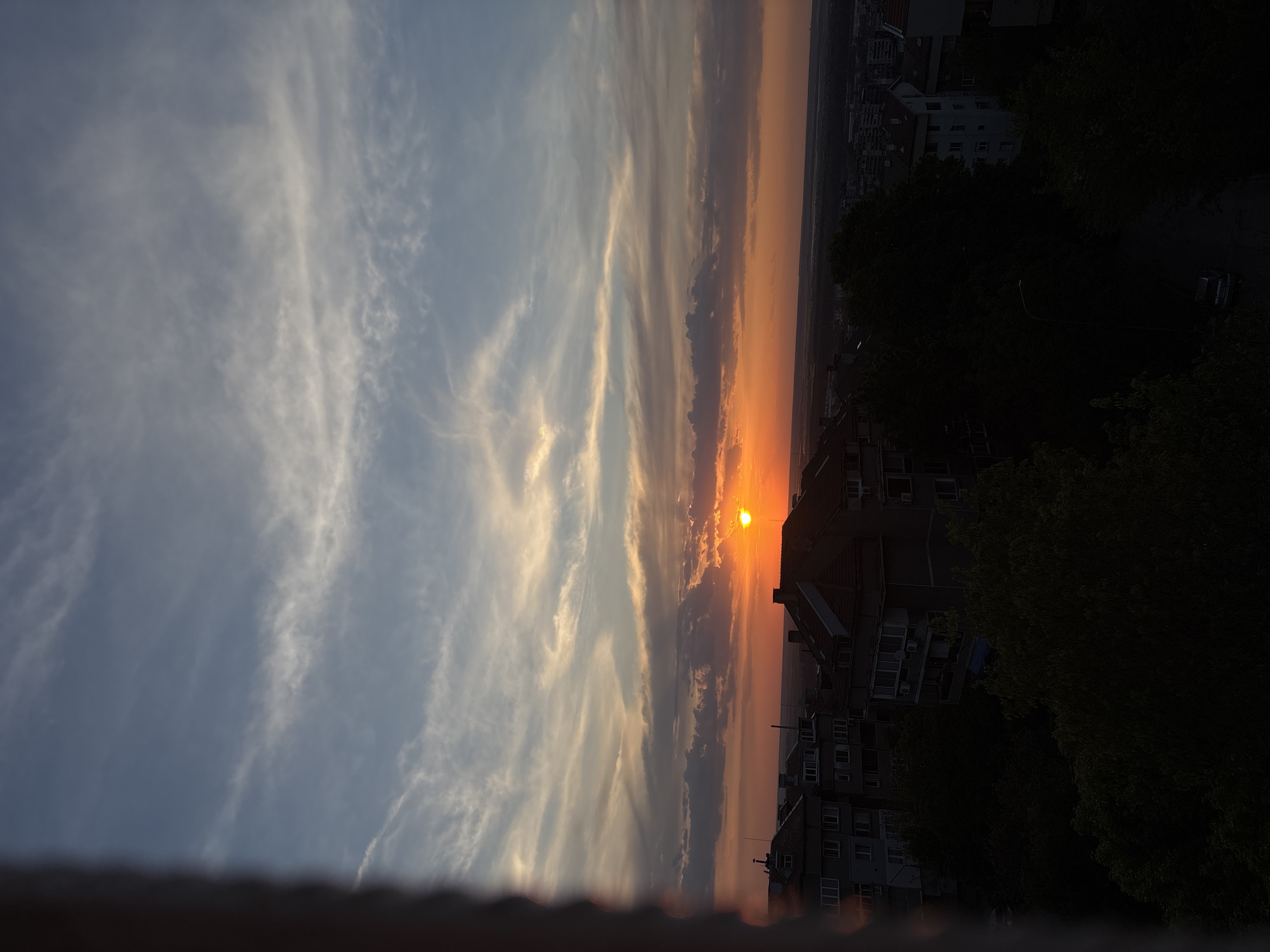 The image shows a sunset viewed from an elevated position in a city. The sky is split into two distinct sections: the left part with a blue sky and scattered white clouds, and the right with orange and darker, elongated clouds closer to the horizon where the sun is setting. The silhouette of buildings, trees, and possibly roads can be seen obscuring the ground and foreground. The sun is positioned low on the horizon, appearing as a bright orange orb.