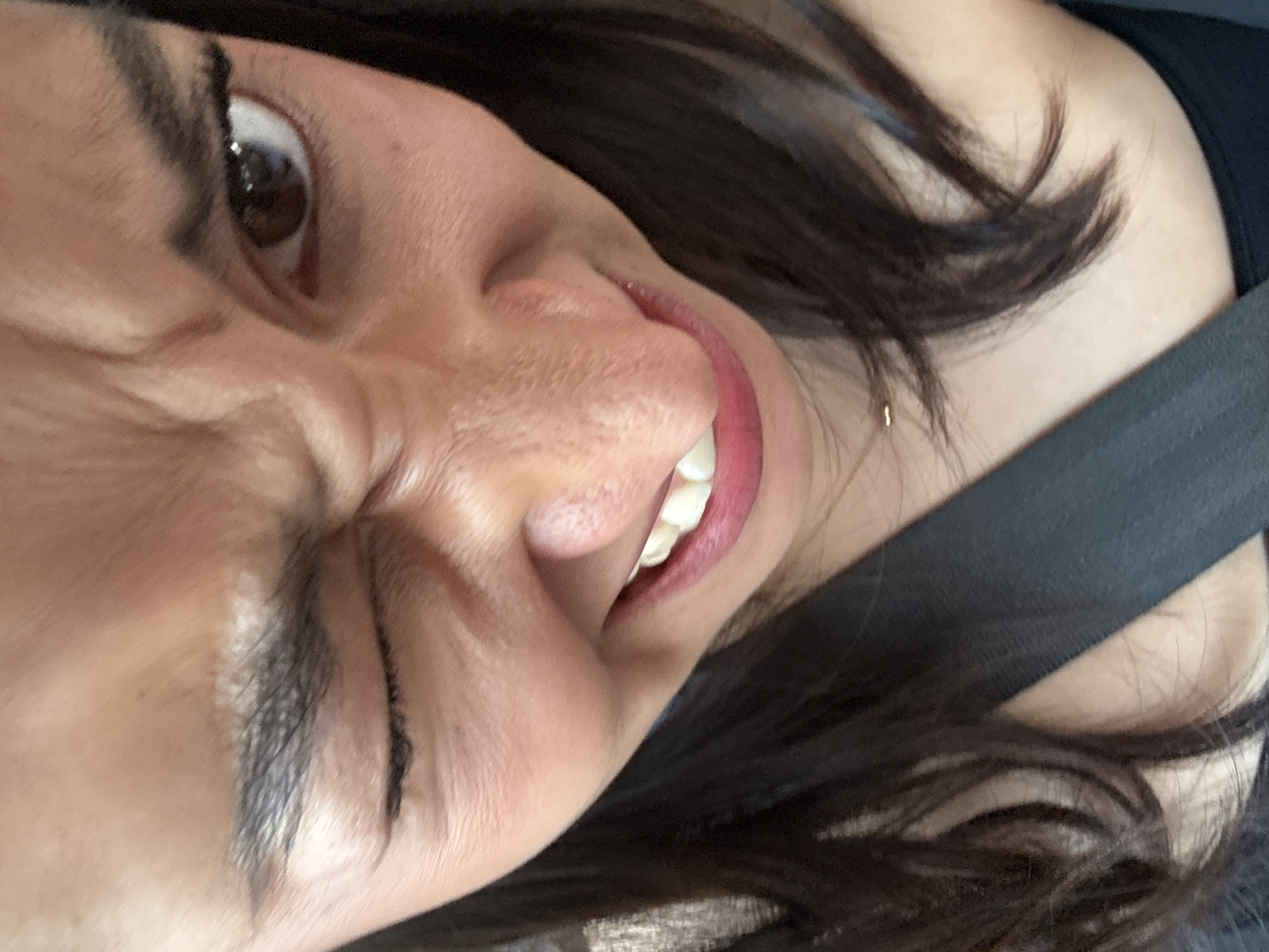 A close-up, rotated image shows a woman's face with her features contorted into a grimace or expression of discomfort. Her left eye is wide open with visible brown iris and pupil, while the surrounding skin is creased. Dark eyebrows and eyelashes are evident. Her nose is wrinkled, and her mouth is slightly open revealing white teeth. She has a gold earring on her neck. Dark hair frames the side of her face. A dark, possibly black, seatbelt strap is visible across her shoulder. The image does not provide sufficient information to determine the location in Puebla, Mexico.