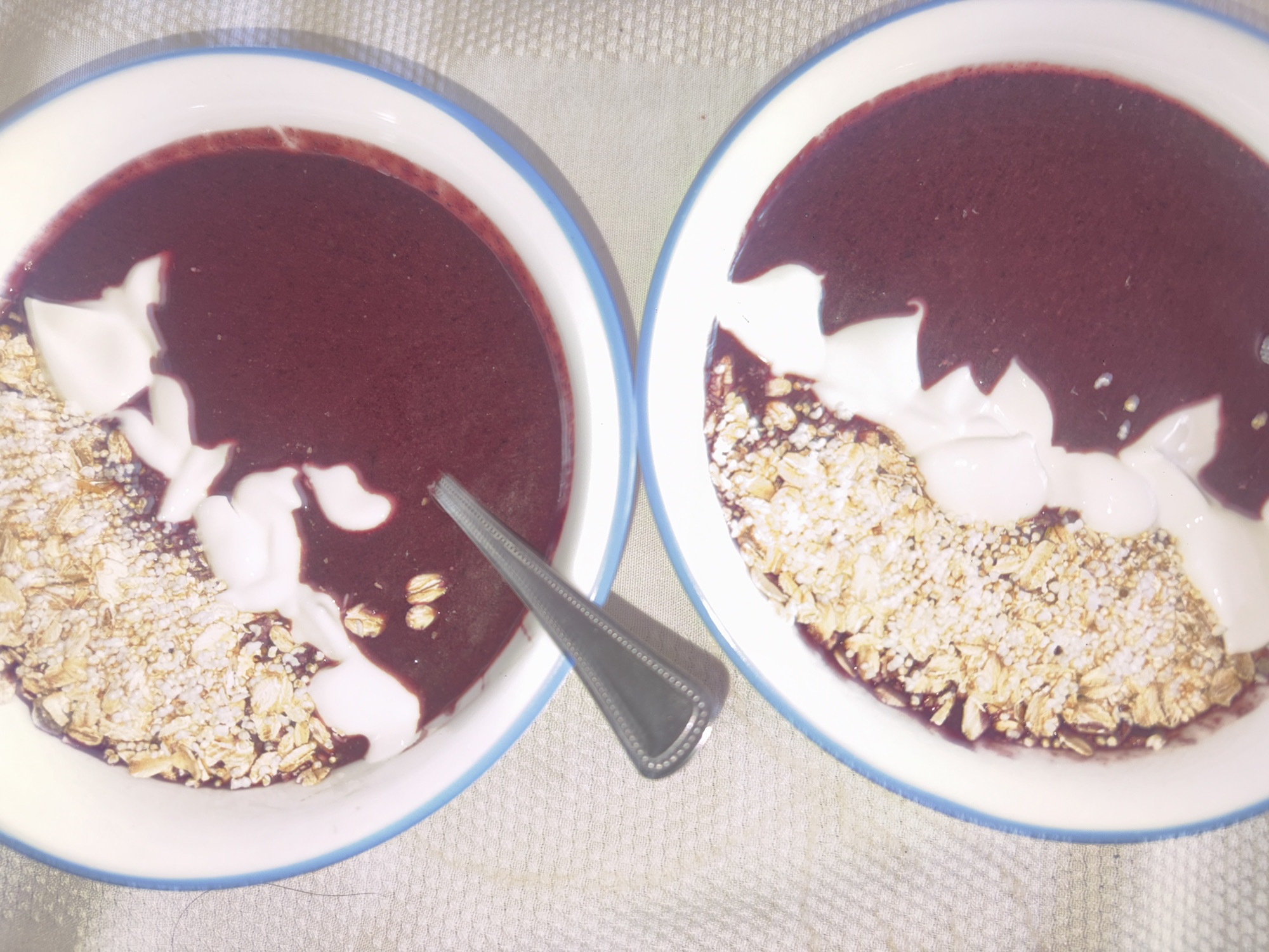 Two bowls of food are side-by-side on a white, textured surface. Each bowl has a white rim with a blue line around the edge. The food in each bowl consists of a dark red liquid base with light brown granola and white dollops on top. A silver spoon lies between the bowls. The location is likely indoors on a countertop or table, but there is no indication that the image was taken in Mexico City, Mexico.