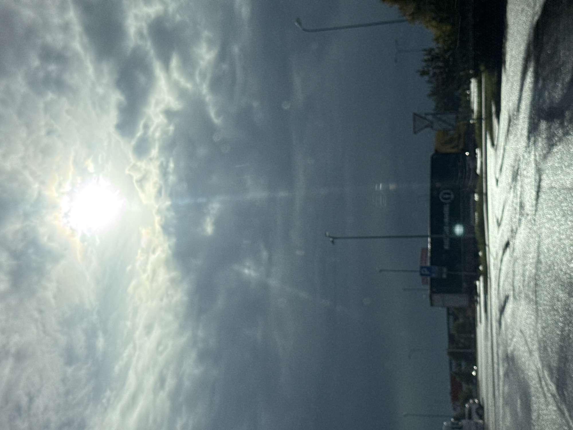 The image depicts an overcast sky with the sun shining through the clouds, creating visible sunbeams. The scene appears to be an outdoor area, possibly a parking lot or industrial zone, with several vehicles including trucks and cars visible. The ground is wet, suggesting recent rain or damp conditions, with tire marks visible on the surface. Streetlights and a traffic sign are also present. Buildings and trees form the background. The overall impression is a somewhat somber or dramatic atmosphere due to the lighting and weather conditions. Based on the visual cues, such as the architecture and general environment, the location is likely an industrial or commercial area on the outskirts of Tallinn, Estonia.