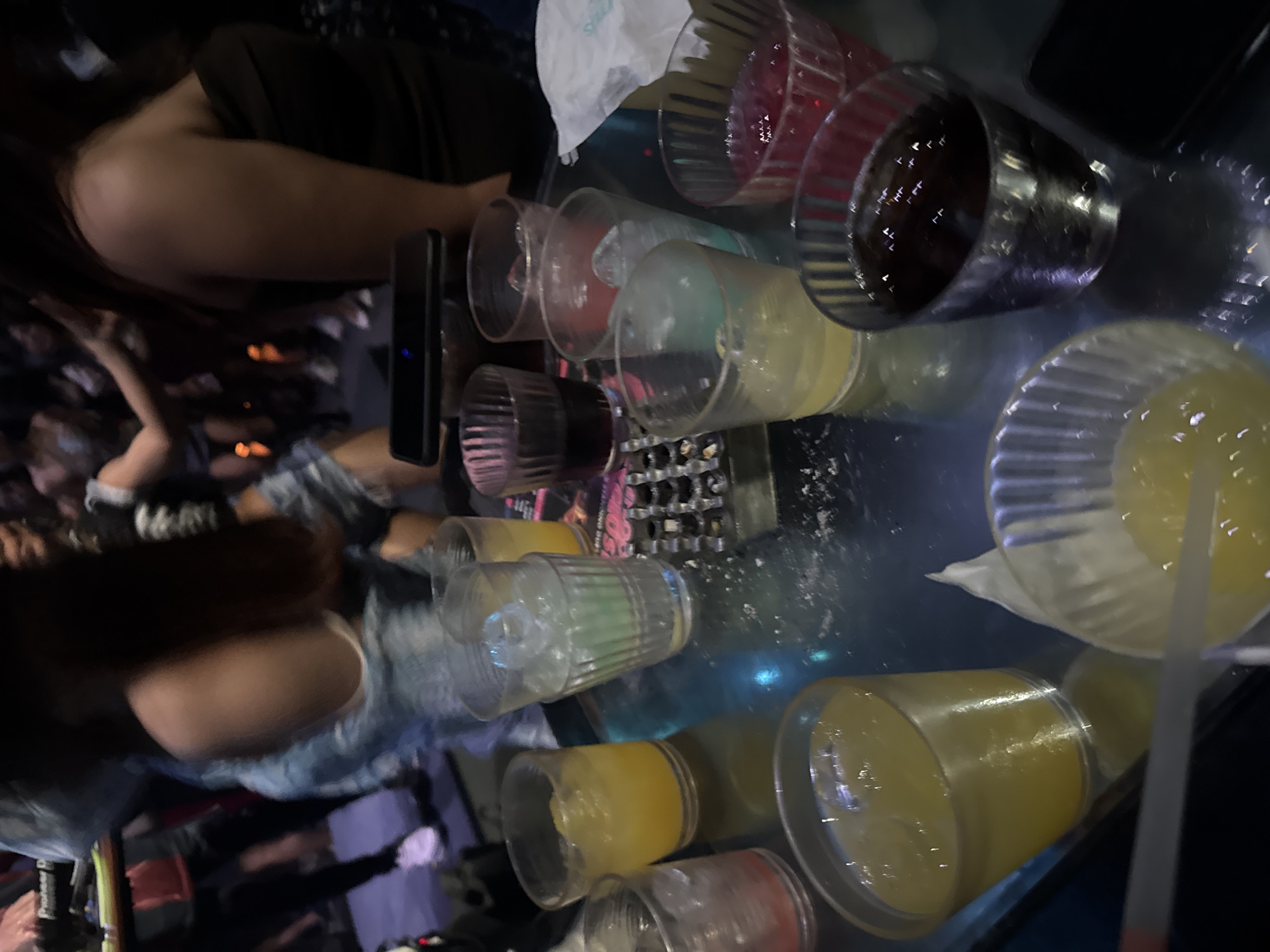 The image depicts a gathering of people at what appears to be a bar or club in North Kuta, Indonesia. Several individuals are visible, though their faces are not clearly discernible due to the low lighting and the orientation of the photo, which is tilted. The primary focus is on a table laden with drinks in various glasses. These include red, yellow, and clear liquids, some with ice. The setting suggests a social event, possibly a night out, given the dim lighting and the presence of multiple drinks. The mood seems to be relaxed and convivial, as implied by the social setting. The people in the image are dressed casually, further suggesting an informal gathering. The reflections on the table surface indicate a glossy material, likely glass or a polished countertop. The time of day is likely evening or night due to the low light conditions. There are no discernible weather cues. No visible text is clearly legible in the image.