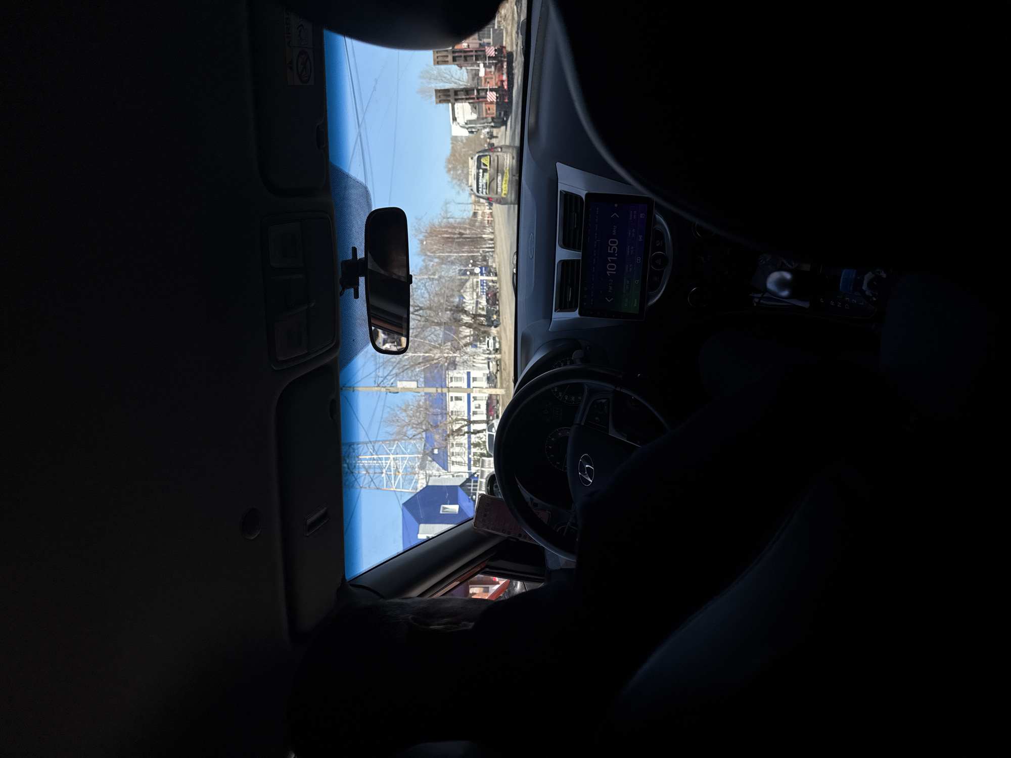 A view from inside a car, presumably the driver's perspective, looking out onto a street in Chaikovsky, Russia. The radio display reads "PMTZ 101.50 MHz." Several buildings and vehicles are visible on the street. The driver is in the car, although their face and torso are partially obscured by shadow.
