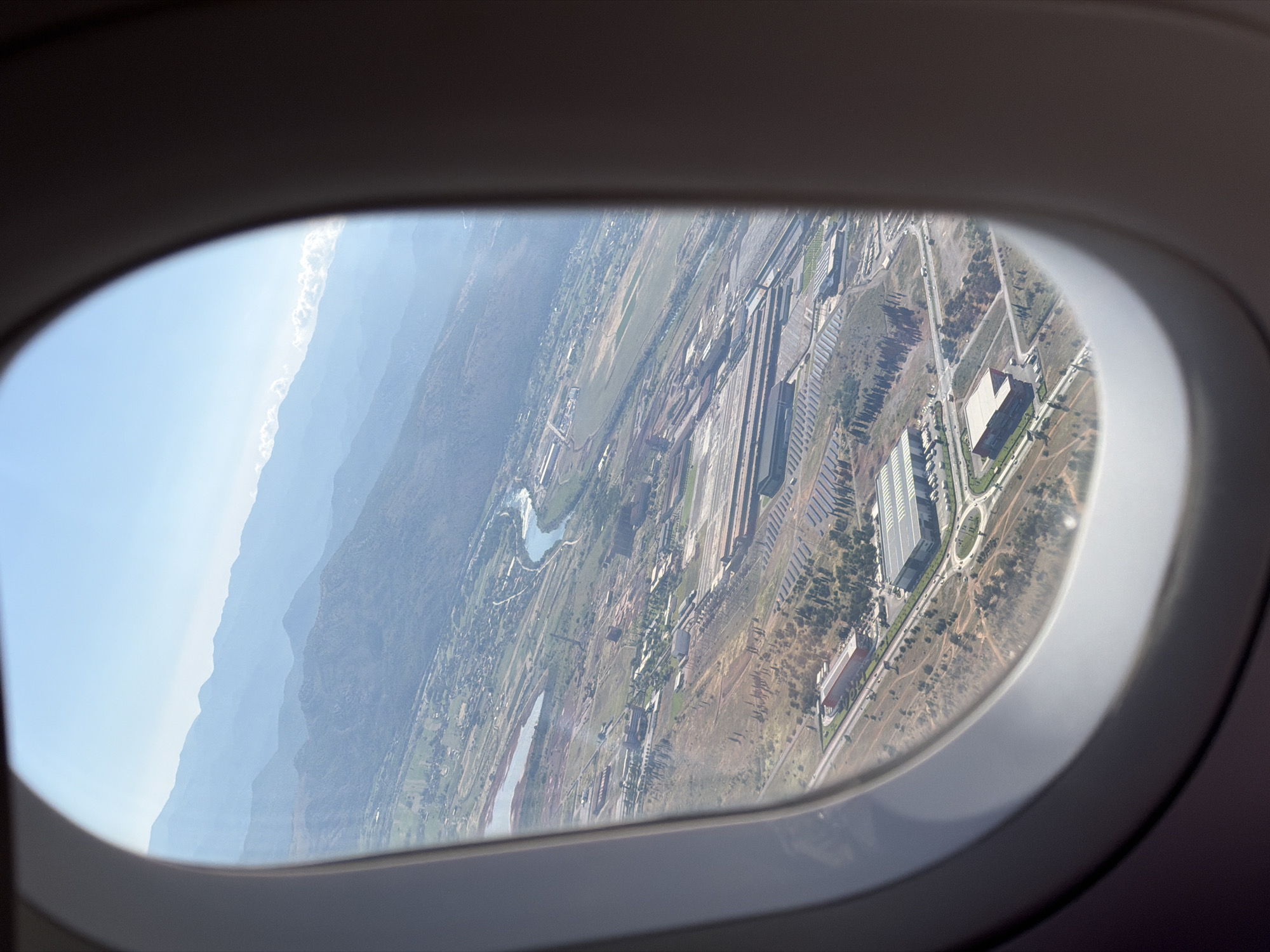 An aerial view from an airplane window shows a landscape with mountains in the distance and an industrial area below. The industrial area appears to be a large complex with multiple buildings, some with large, flat roofs, and what could be solar panels arranged in rows. A river or body of water snakes through the landscape, and roads with some vehicles are visible. The terrain is uneven, with some areas of sparse vegetation and rocky soil. The sky is clear and blue, with distant mountains fading into a hazy horizon. The image captures a broad view of the environment, focusing on the sprawling industrial development within a mountainous region.