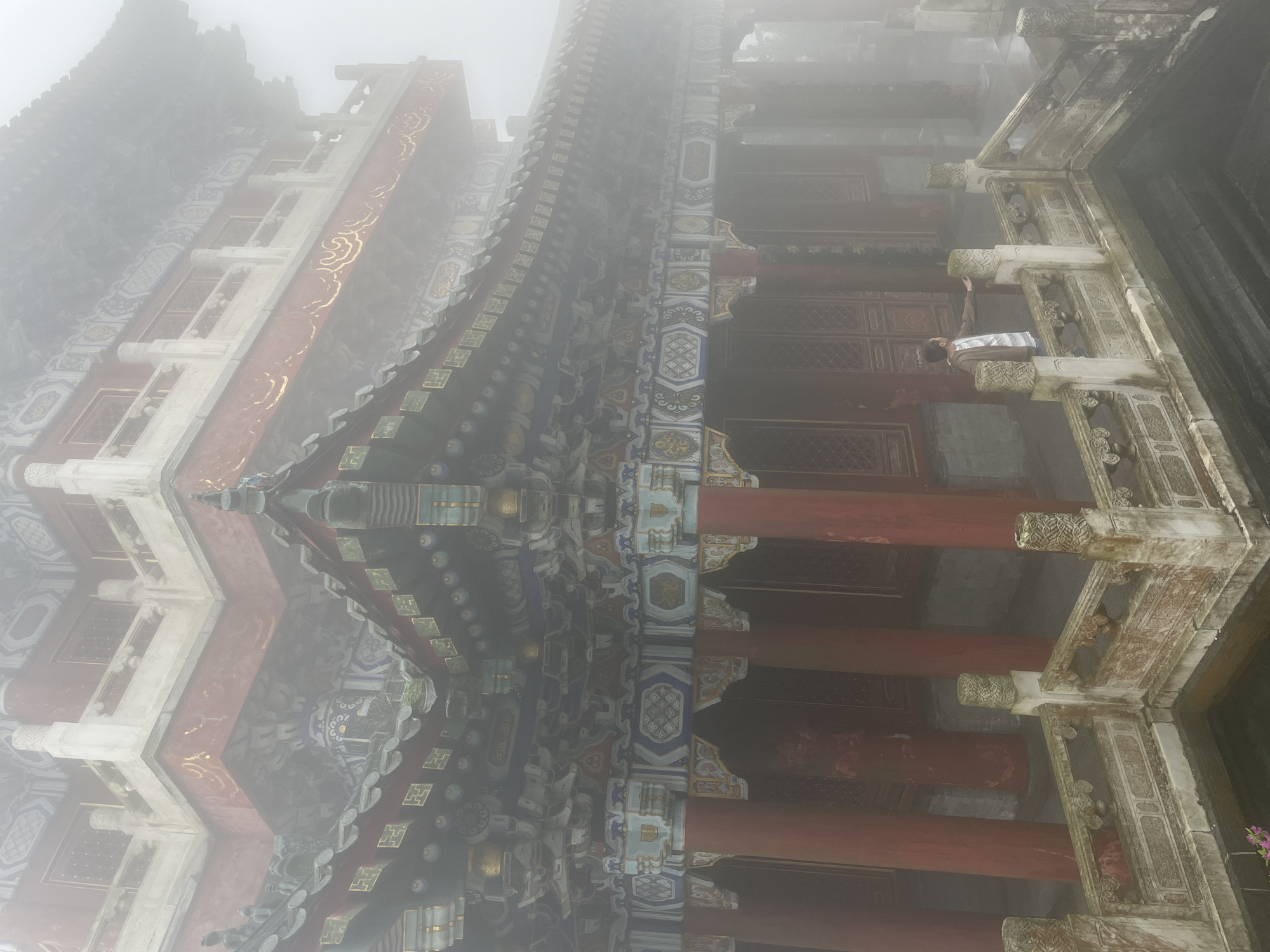 A low-angle, hazy photograph captures a section of an ornate, traditional Chinese building. The architecture features red columns supporting intricately decorated eaves in shades of blue, gold, and white. Visible through an arched opening, a person stands partially concealed, their arm reaching towards the structure. The scene is enveloped in fog, obscuring details and creating a muted color palette. The location is likely within Yongding District, China.