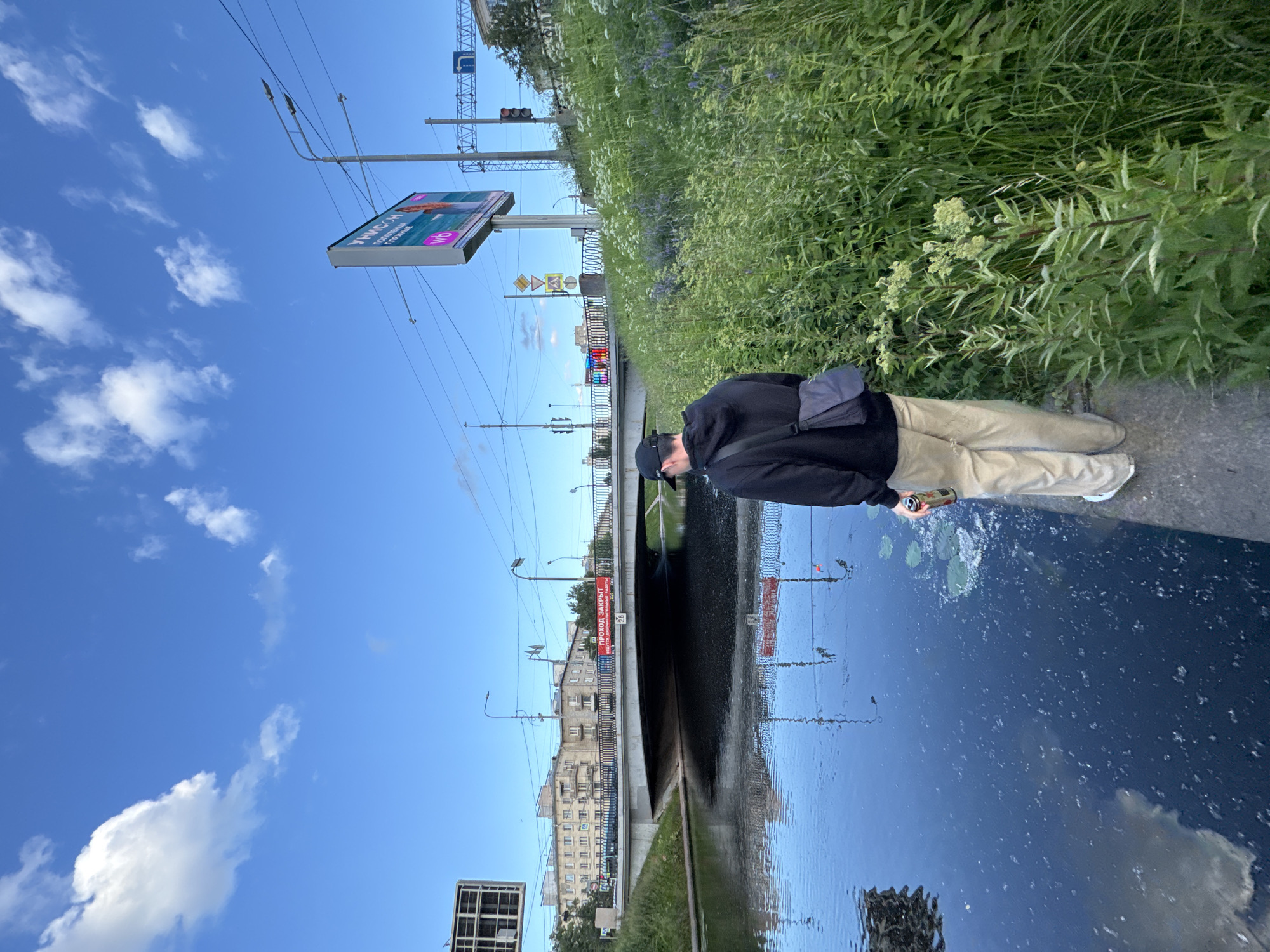The image depicts a man standing by the Krestovka River in Primorsky District, Saint Petersburg, Russia. The man, wearing a black hoodie, beige pants, a black baseball cap, and a gray crossbody bag, holds a can of beer. He is facing away from the camera, looking at the river. The river water is dark and reflects the sky and bridge. Lush green vegetation lines the riverbank, with some purple flowers visible. In the background, a concrete bridge spans the river, with apartment buildings and a billboard visible on the other side. The sky is blue with scattered white clouds.