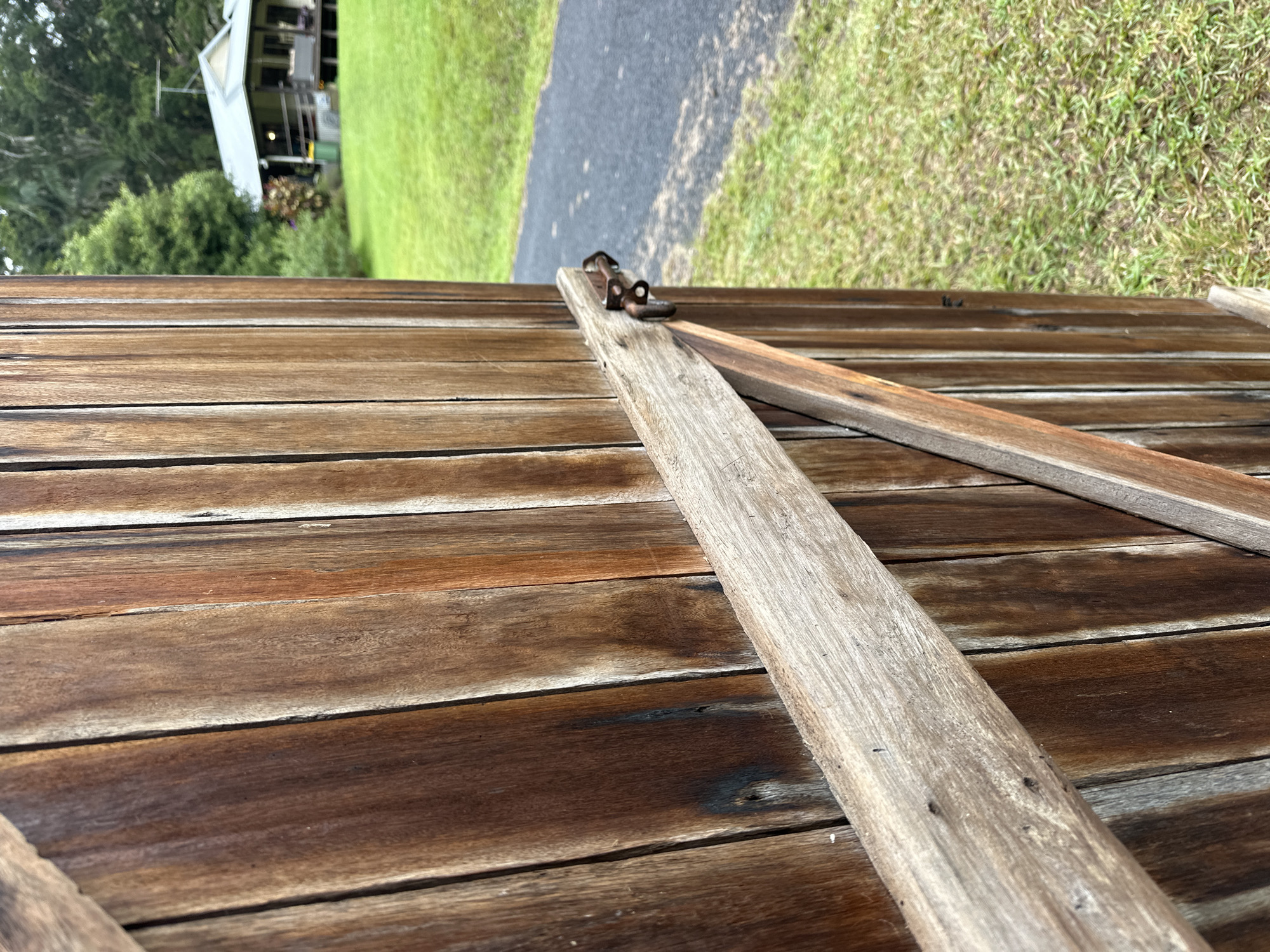 The image is a close-up, eye-level, outdoor shot of a weathered wooden gate. The gate is made of vertical planks of dark brown and reddish-brown wood, with visible grain and some dark discoloration. A diagonal wooden support beam is attached to the gate, and a rusty metal latch mechanism is visible securing it. To the right of the gate, a paved pathway runs through a grassy area. In the background, a house with a grey roof and a wide porch is visible, surrounded by lush green trees and foliage. The scene appears to be a rural or semi-rural setting. The metadata indicates the location is Thora, Australia.