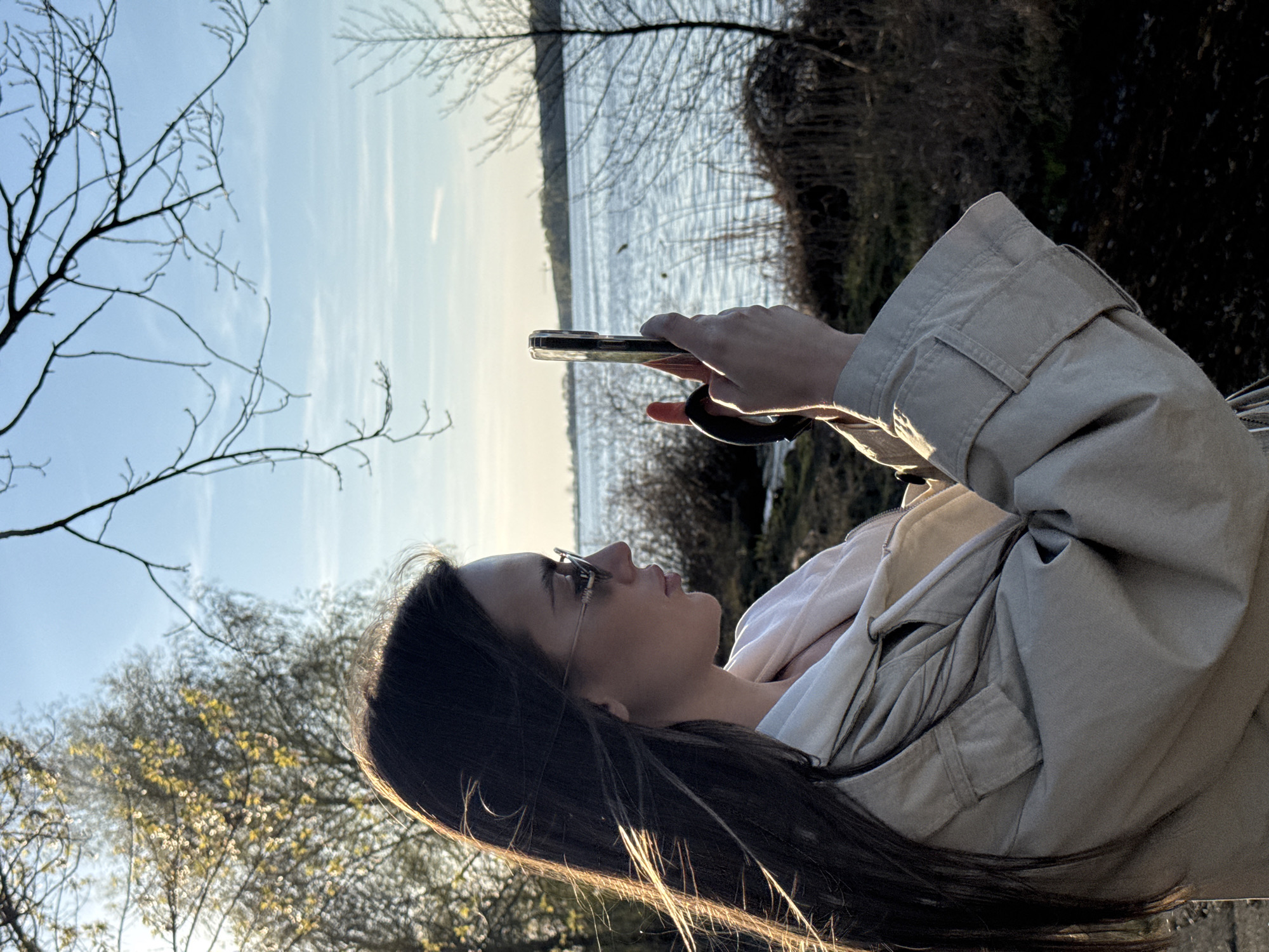 A woman is seen in profile, holding a mobile phone up as if taking a photo or video. She has long brown hair and is wearing glasses and a light-colored jacket over a lighter shirt. The background shows a body of water with trees lining the opposite bank. Some trees are also visible in the foreground, appearing bare or with early spring buds. The sky is light blue with some wispy clouds. Considering the landscape, this scene is likely set near the Pilica River, a common feature of Białobrzegi, Poland. The location could be near the riverbanks or a recreational area along the Pilica, often used for nature walks and photography. The woman is likely capturing the scenic view.