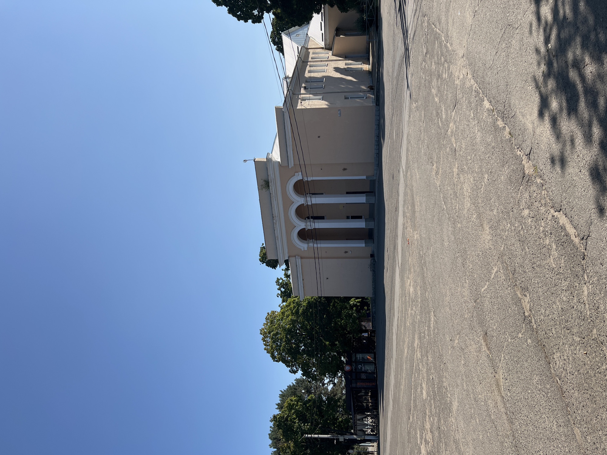A wide, empty paved area stretches out in the foreground under a clear blue sky. In the background, a light peach-colored building with arched entrances and white columns stands prominently. To the left of the building, a dark-colored structure with awnings and signage is visible, along with several mature green trees. The paving in the foreground shows signs of wear and tear, with visible cracks and patches. The scene appears to be a public square or plaza in front of a civic building, likely in Dubăsari town council, Moldova.