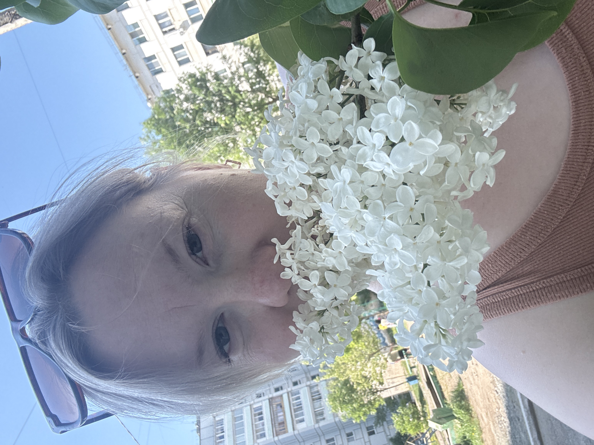 A woman with fair skin and light hair poses close to the camera, holding a large bouquet of white flowers to her face. Her eyes are visible, and a pair of sunglasses is perched on her head. She wears a light brown, textured top. The bouquet partially obscures her face.
In the background, an urban landscape is visible, including a light-colored building with multiple windows. Green trees and a grassy area with benches are also visible, suggesting a park or residential area. The setting appears to be outdoors in daylight. The location is Chaikovsky, Russia.