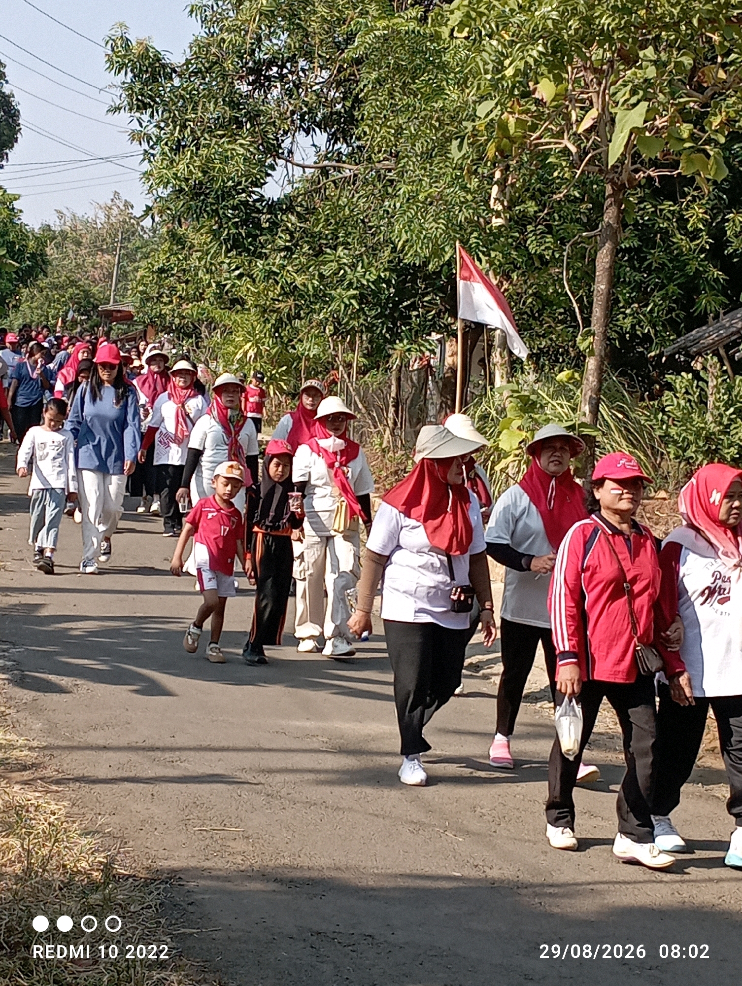 A group of people, including adults and children, are walking down a paved road lined with trees and vegetation. Many of the individuals are wearing red and white attire, and some have headscarves or hats. A small Indonesian flag is visible on a pole to the right of the road. The sun is shining, casting shadows on the road. The image appears to be taken in Dukuhseti, Indonesia, as indicated by the timestamp "REDMI 10 2022" and "29/08/2026 08:02". The people are walking in the same direction, suggesting a group activity or procession. Some individuals are holding small bags or cups. The environment is rural, with foliage and a natural setting.