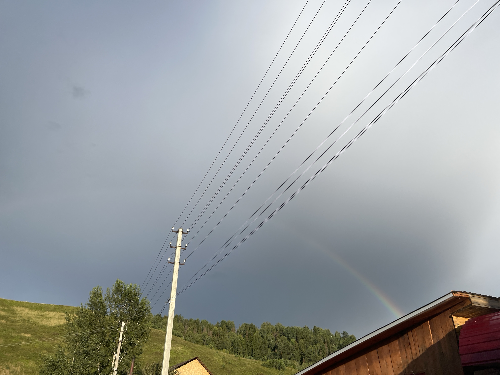 The image shows a rural landscape under a cloudy sky. In the foreground, a wooden building with a red roof is partially visible on the right. A power pole with multiple wires stretches across the upper portion of the frame. To the left of the power pole, a green grassy hillside rises, dotted with trees and a dense forest line in the background. A faint rainbow is visible in the sky towards the right side of the image, suggesting recent or ongoing rain. The overall scene depicts a natural environment with man-made elements. The location is identified as Ольховка, Russia. There are no people or active interactions depicted in the image.