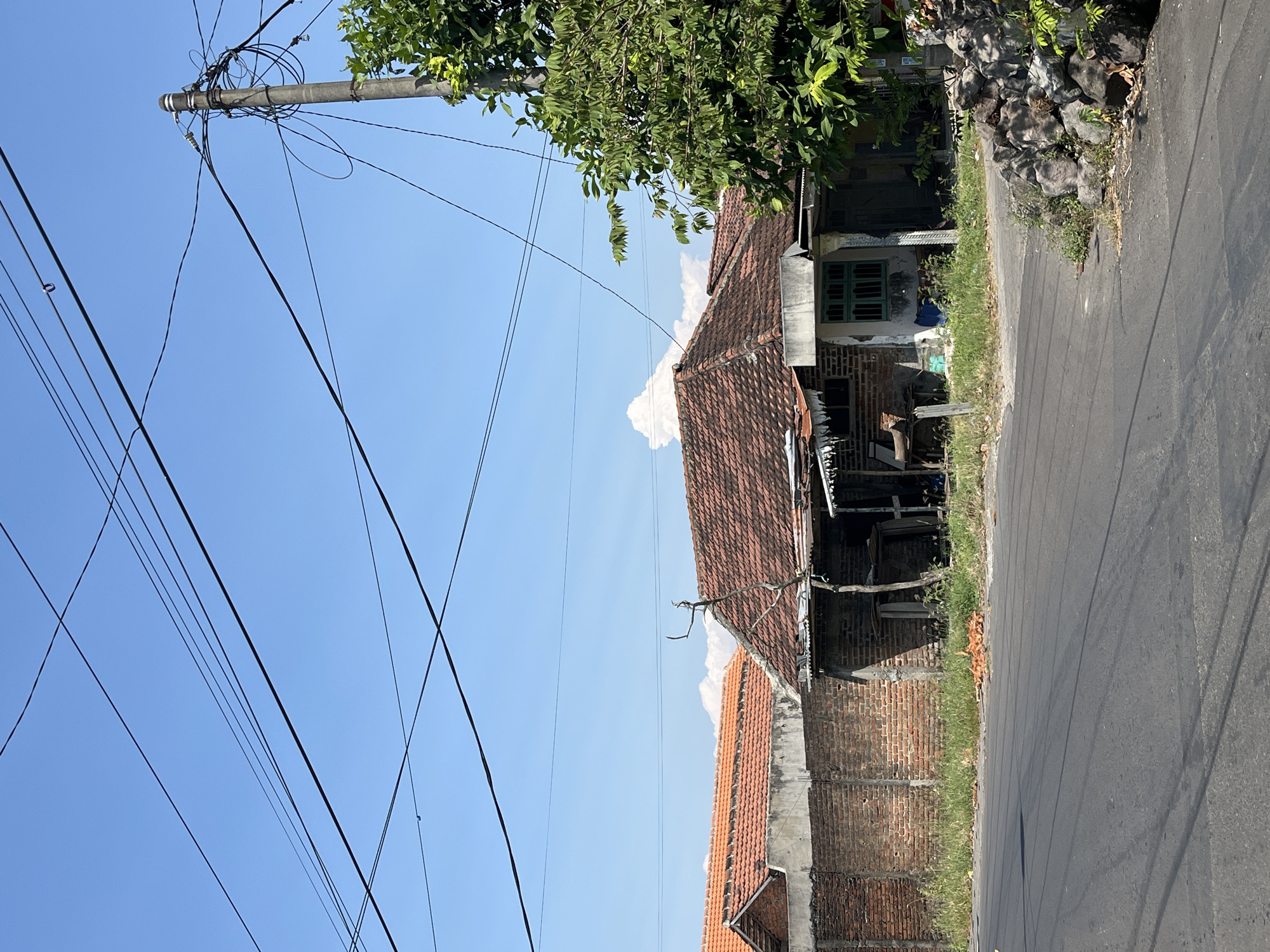 The image shows a street scene in the city of Pesawahan, Indonesia. In the foreground, a paved road runs diagonally from the bottom left to the middle right of the frame. The road has visible tire marks and is bordered by a patch of dry grass and some scattered rocks on the right.

Behind the road, there are several structures. A prominent building with a red tiled roof and exposed brick walls sits in the middle ground, to the right of the frame. This building appears to be a house or a small shop, with an open front area supported by wooden posts. Adjacent to it, to the left, is another building with a similar red tiled roof, separated by a brick wall. This building also has brick walls. The area between these buildings and the road is covered in overgrown grass and some debris.

In the background, the sky is clear and blue with a few wispy white clouds. Overhead, a network of black electrical wires crisscrosses the sky, supported by a concrete utility pole on the right side of the image. The utility pole is laden with various wires and equipment. A large green leafy tree is situated to the right of the utility pole, partially obscuring the sky. The overall impression is a simple, rural or semi-urban street in a tropical climate.