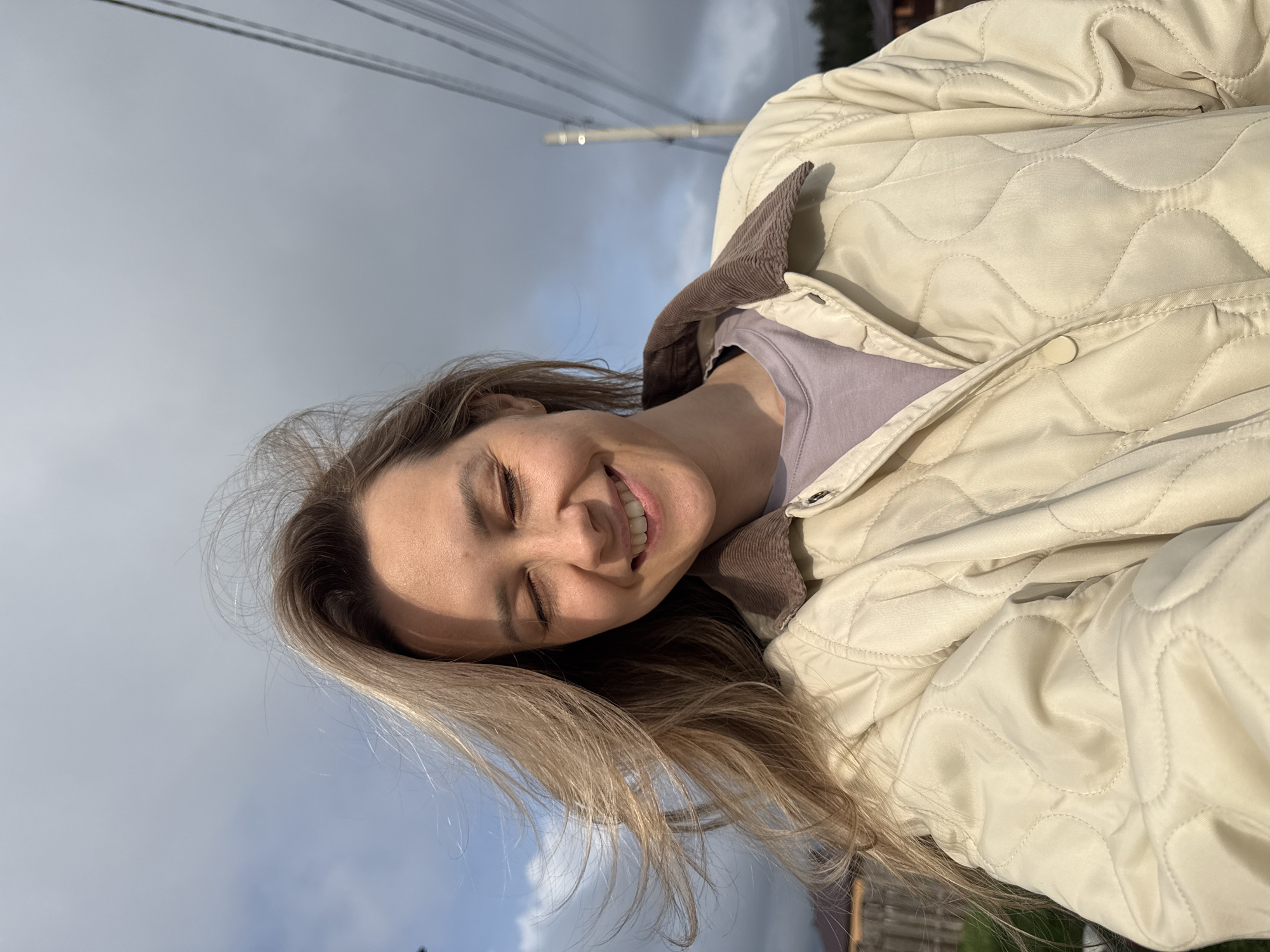 A woman with long blonde hair is smiling with her eyes closed. She is wearing a light beige quilted jacket with a brown corduroy collar over a lavender t-shirt. The background is a cloudy sky with utility poles and wires visible. Part of a wooden fence and some greenery can be seen in the lower right corner. This photo was taken outside in a location in Chaikovsky, Russia.