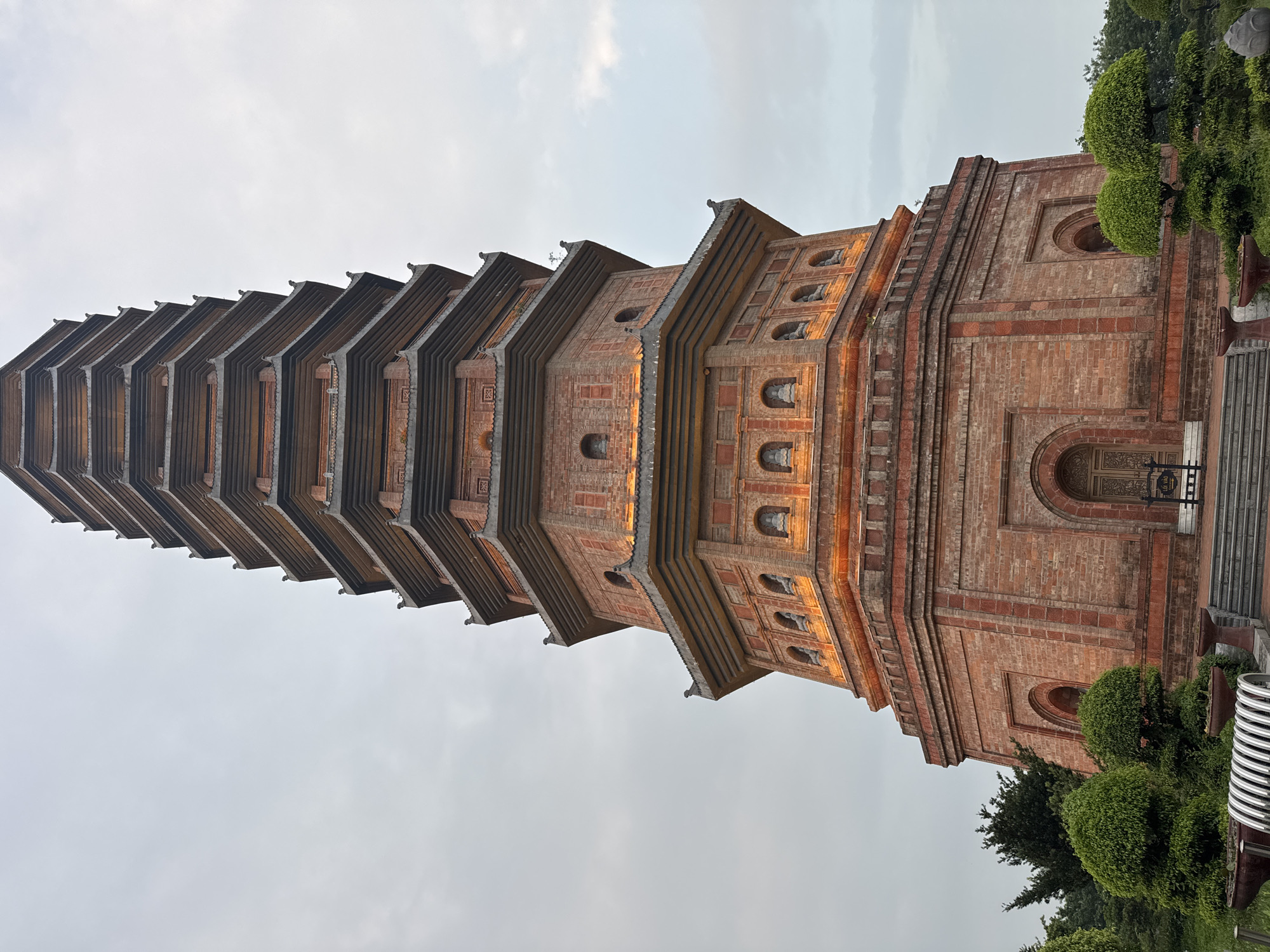 This image depicts the 9-story Bai Dinh Pagoda, a prominent Buddhist temple complex located in the commune of Gia Sinh, Gia Vien district, Ninh Binh province, near the city of Tây Hoa Lư Ward, Vietnam. The pagoda is constructed of brick and features multiple tiers, each with curved, tiled roofs characteristic of traditional Vietnamese architecture. Niches containing Buddha statues are visible on several levels, illuminated by warm orange lighting. The pagoda's base is surrounded by manicured greenery, including intricately shaped bushes and trees, as well as planters and seating. The sky above is overcast, suggesting diffused daylight or evening conditions. The foreground shows a pathway leading to the pagoda entrance, with steps and decorative elements.
