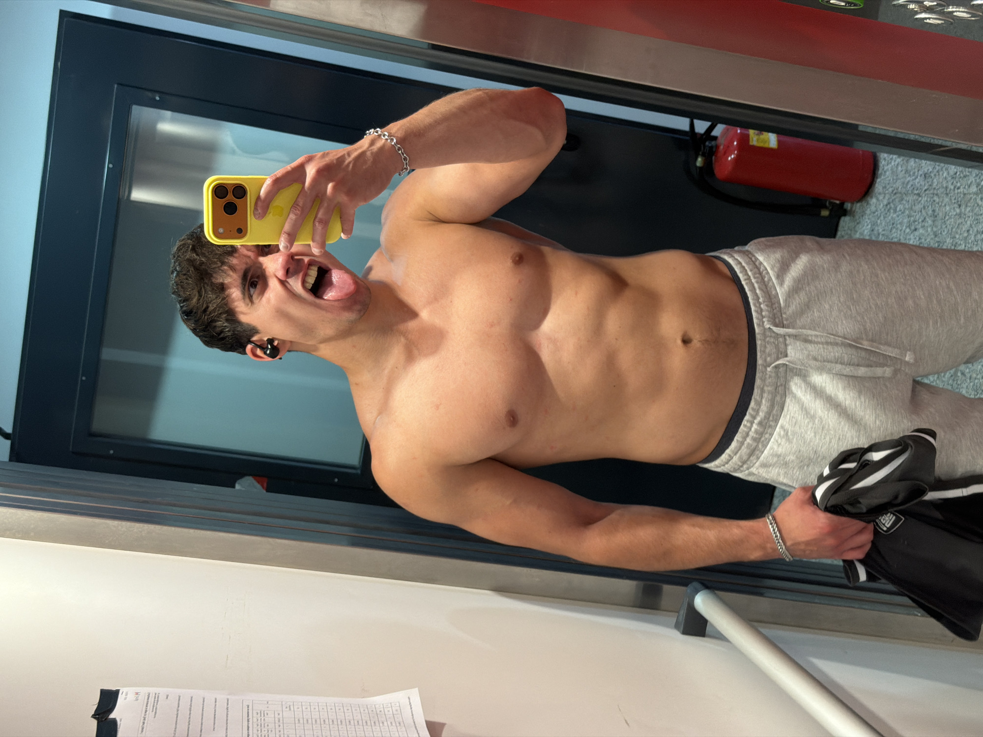 A young man with a muscular physique is depicted in what appears to be a mirror selfie, seen from the waist up. He is shirtless, showcasing his developed torso and abs, and wears light grey sweatpants with a dark elastic waistband. His dark hair is somewhat disheveled, and he has a playful, energetic expression with his mouth wide open, tongue sticking out, and eyes wide. He holds a bright yellow smartphone with a visible triple camera setup in his right hand, pointed towards himself. A black earbud is visible in his left ear, and a silver chain bracelet adorns his left wrist.
In his left hand, he holds a folded black garment with white stripes. The background suggests an indoor location, possibly a gym or sports facility, with a dark-framed window or door on the left. A red fire extinguisher is mounted on the wall to the right. A silver bar or railing extends across the lower part of the frame, and papers with unreadable text are visible on a light surface at the bottom left. The image conveys a confident and dynamic mood. The scene is located in Sofia, Bulgaria.