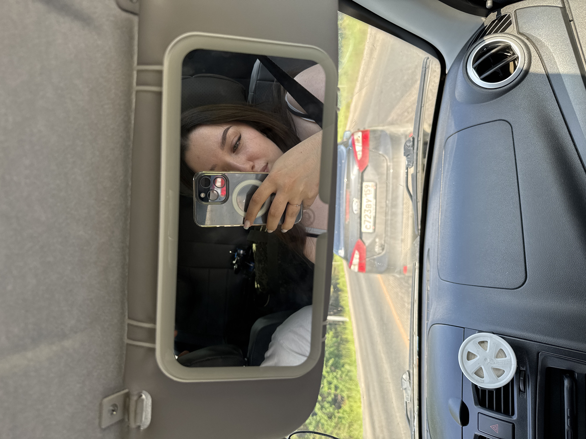 The image shows a young woman taking a selfie in a car's vanity mirror. She is wearing a light-colored tank top and has long, dark hair. She is holding a smartphone in her right hand, which is reflected in the mirror. Her left hand, adorned with a ring, is also visible. In the background of the mirror's reflection, another person wearing a light-colored t-shirt is visible in the passenger seat. The car appears to be in motion, as indicated by the road and other vehicles visible through the windshield. A dark-colored car with a license plate reading "C723BY 159" is directly in front of them. The interior of the car features a dashboard with an air freshener and a circular air vent. The scene takes place on a sunny day on a paved road, likely in a rural or suburban area of Pesterevo, Russia, given the presence of greenery and trees along the roadside.