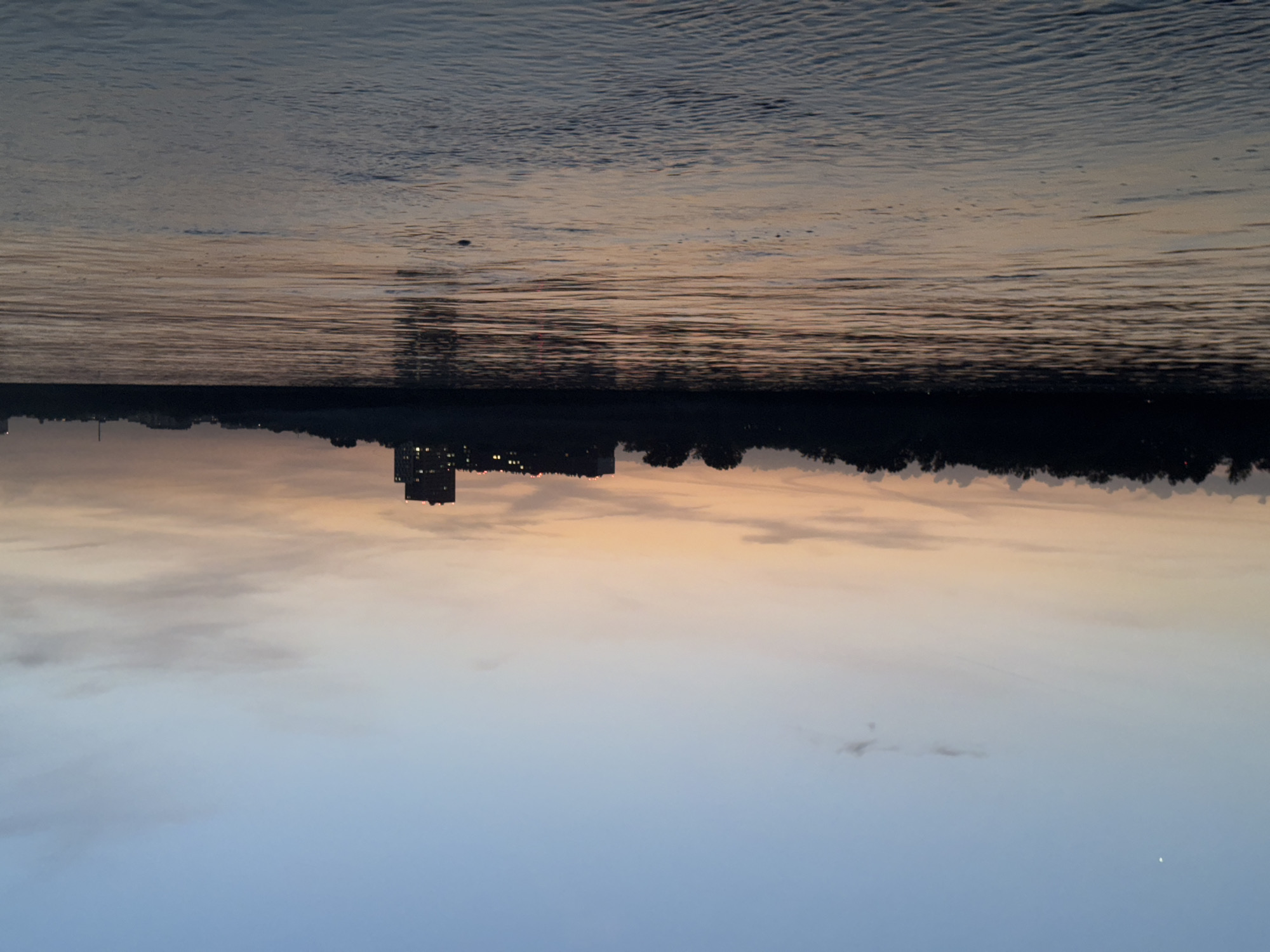 The image captures a scene at dusk or dawn, with the sky transitioning from a light blue at the bottom to orange and purple hues near the horizon. A body of water, likely a river or lake, occupies the upper portion of the image, reflecting the colors of the sky. Along the horizon line, there's a dark silhouette of land, which includes a building with illuminated windows, possibly an apartment complex or office building, along with a line of trees. No people are visible in the image. The setting appears to be on the outskirts of a city, where natural elements meet urban development.
