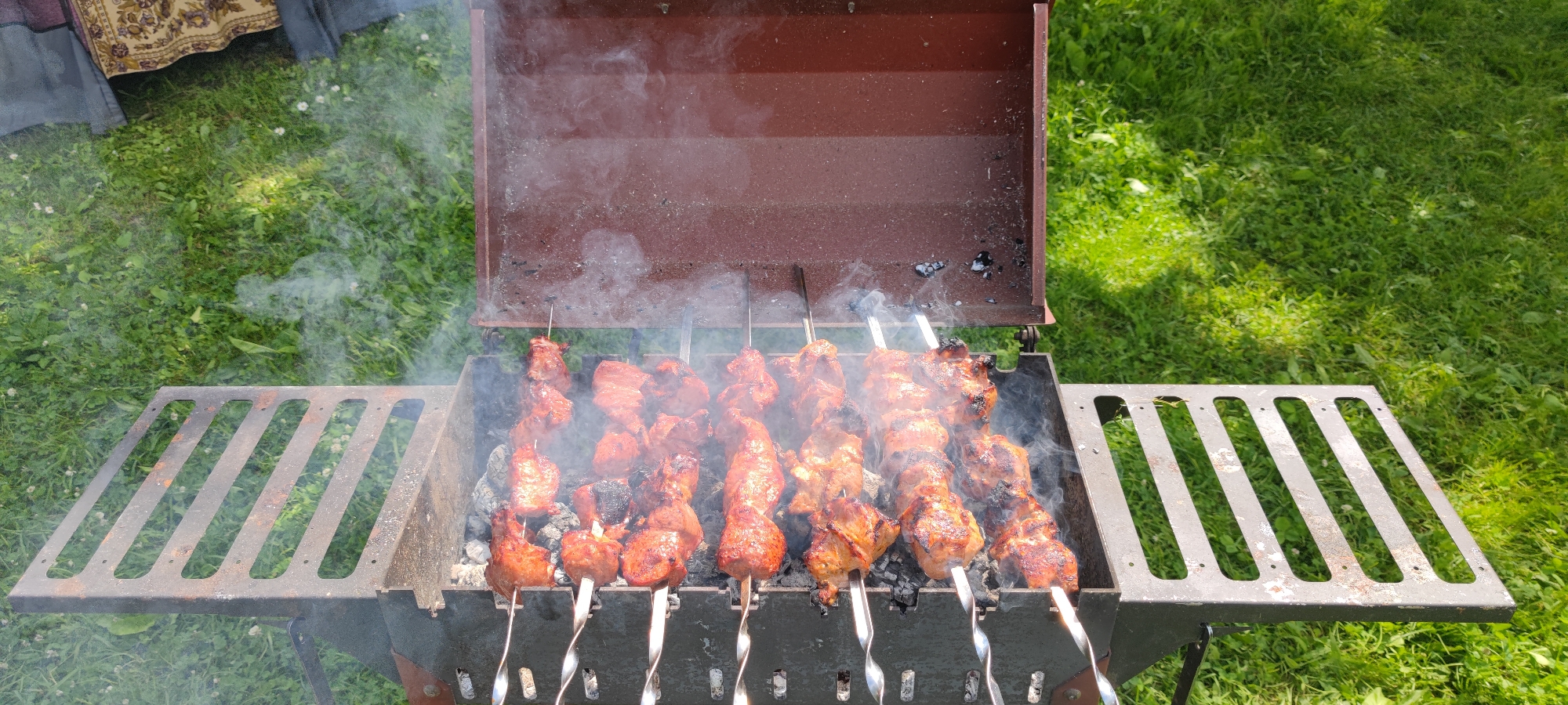 The image captures an outdoor barbecue scene, likely in a rural setting such as Пацейкаўскі сельскі Савет, Belarus, given the lush green grass and natural light. A metal barbecue grill, a common feature for outdoor cooking, is centrally placed. The grill is filled with hot coals, emitting visible plumes of smoke that rise and obscure parts of the food being cooked.
Several skewers, each laden with pieces of meat, are arranged horizontally across the grill, undergoing the cooking process. The meat appears to be seasoned and is in various stages of browning, some pieces showing char marks indicative of grilling. The skewers themselves are made of metal, with spiral-shaped handles at the visible ends, suggesting they are designed for easy turning.
The grill has a hinged lid, which is open and angled upwards, allowing the smoke to escape. The grill is supported by a dark-colored metal frame, and to its left, there's a slatted metal rack extending outwards, possibly for placing food or utensils.
The surrounding environment is dominated by a vibrant green lawn, dotted with small white flowers, consistent with a natural outdoor space. In the upper left corner of the image, a portion of a colorful textile is visible, possibly a tablecloth or a decorative fabric, hinting at a gathering or picnic. The overall impression is one of a casual, enjoyable outdoor meal preparation.