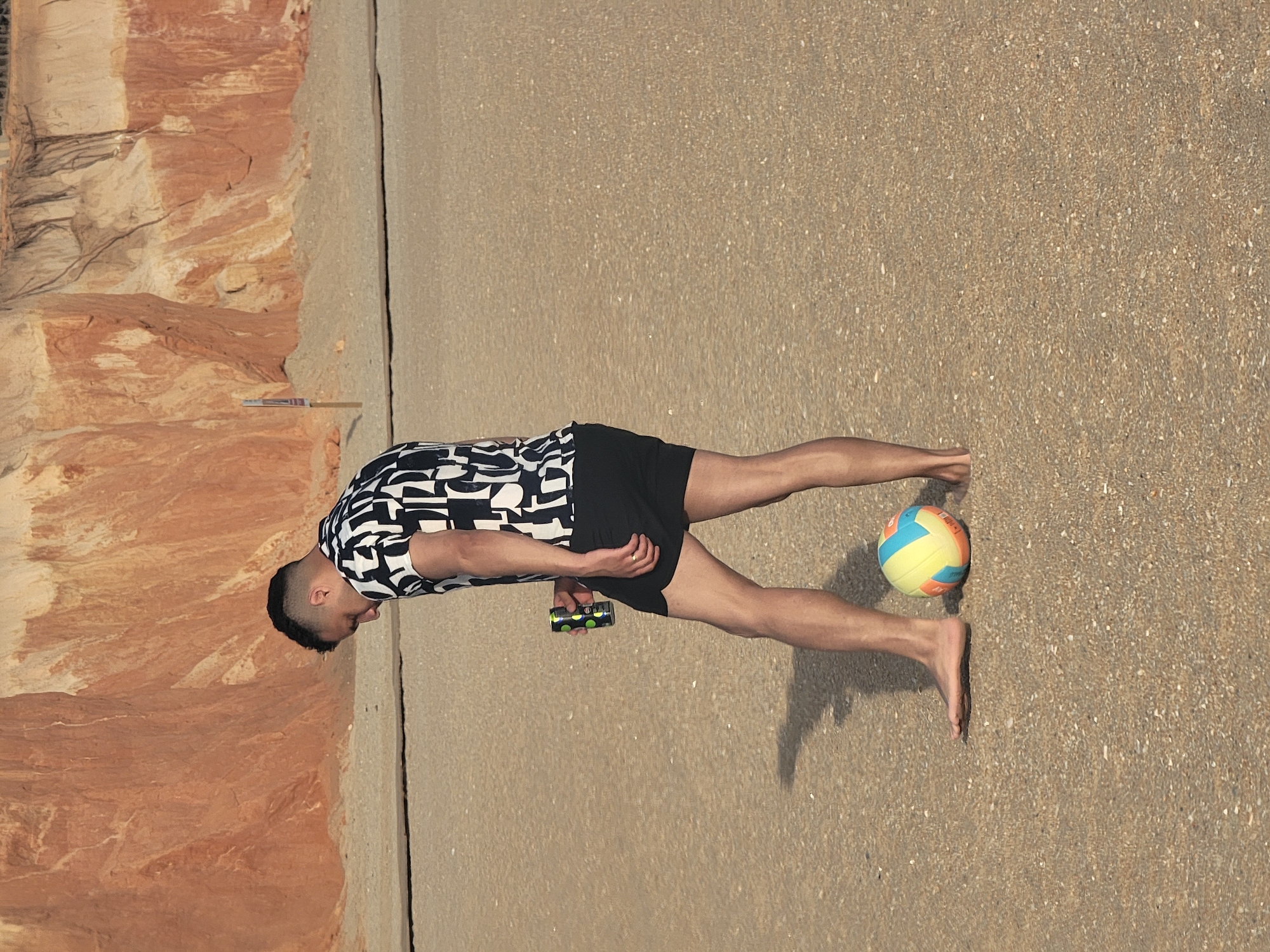 A young man is standing on a sandy beach in Quarteira, Portugal, with a colorful volleyball at his feet. He is wearing a black and white patterned short-sleeved shirt and black shorts. In his right hand, he holds a can with green and black dots. Behind him, a large reddish-brown cliff face is visible, with some debris and vegetation at its base. The beach appears to be sparsely populated, with a wooden boardwalk running horizontally in the background. The lighting suggests it is daytime.