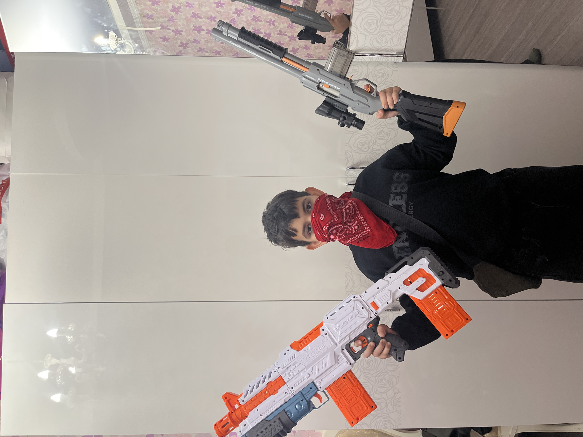 A young boy stands in a room, holding two toy rifles. He is wearing a black hoodie and a red bandana over his face. The room appears to be a bedroom or dressing room, with a large white wardrobe and floral wallpaper. A chandelier is visible in the reflection on the wardrobe doors. The boy is positioned centrally in the frame, with the toy rifles held out in front of him. One rifle is white, orange, and blue, while the other is gray and orange. The image is likely taken indoors, possibly in Kostroma, Russia, based on the provided context.